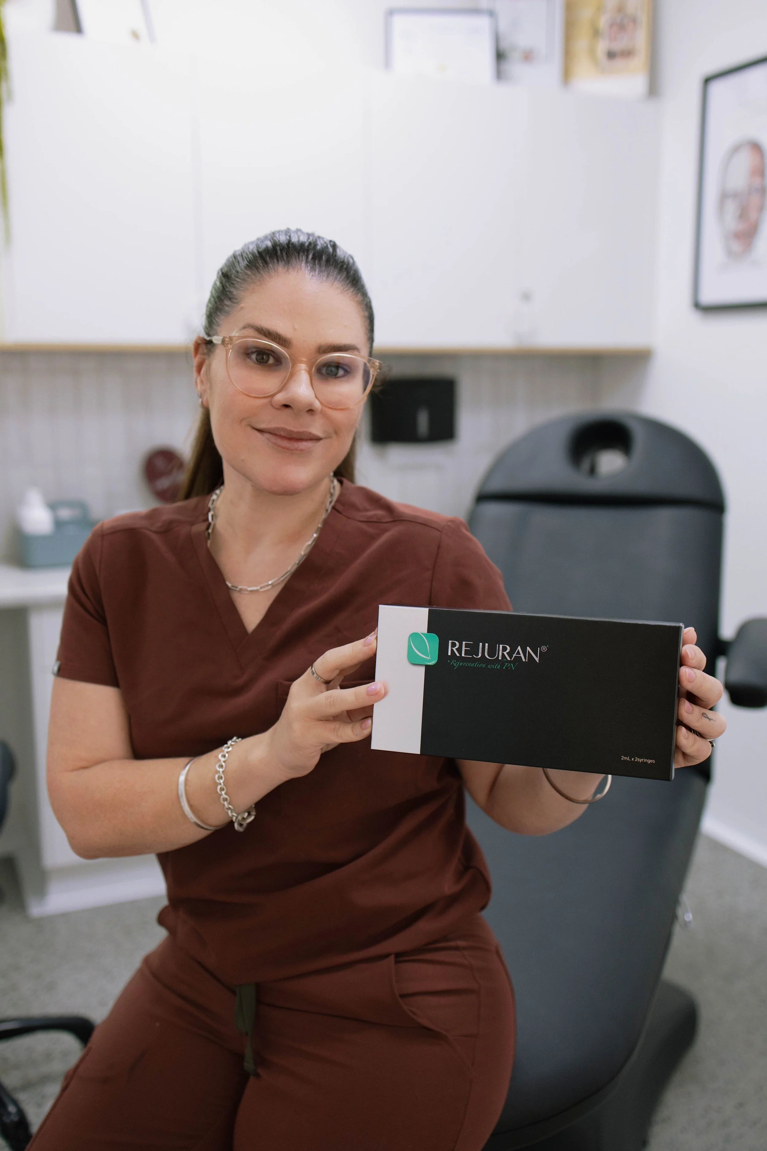 A woman wearing glasses holding a box labeled Rejuran, seated in an office or clinic setting.