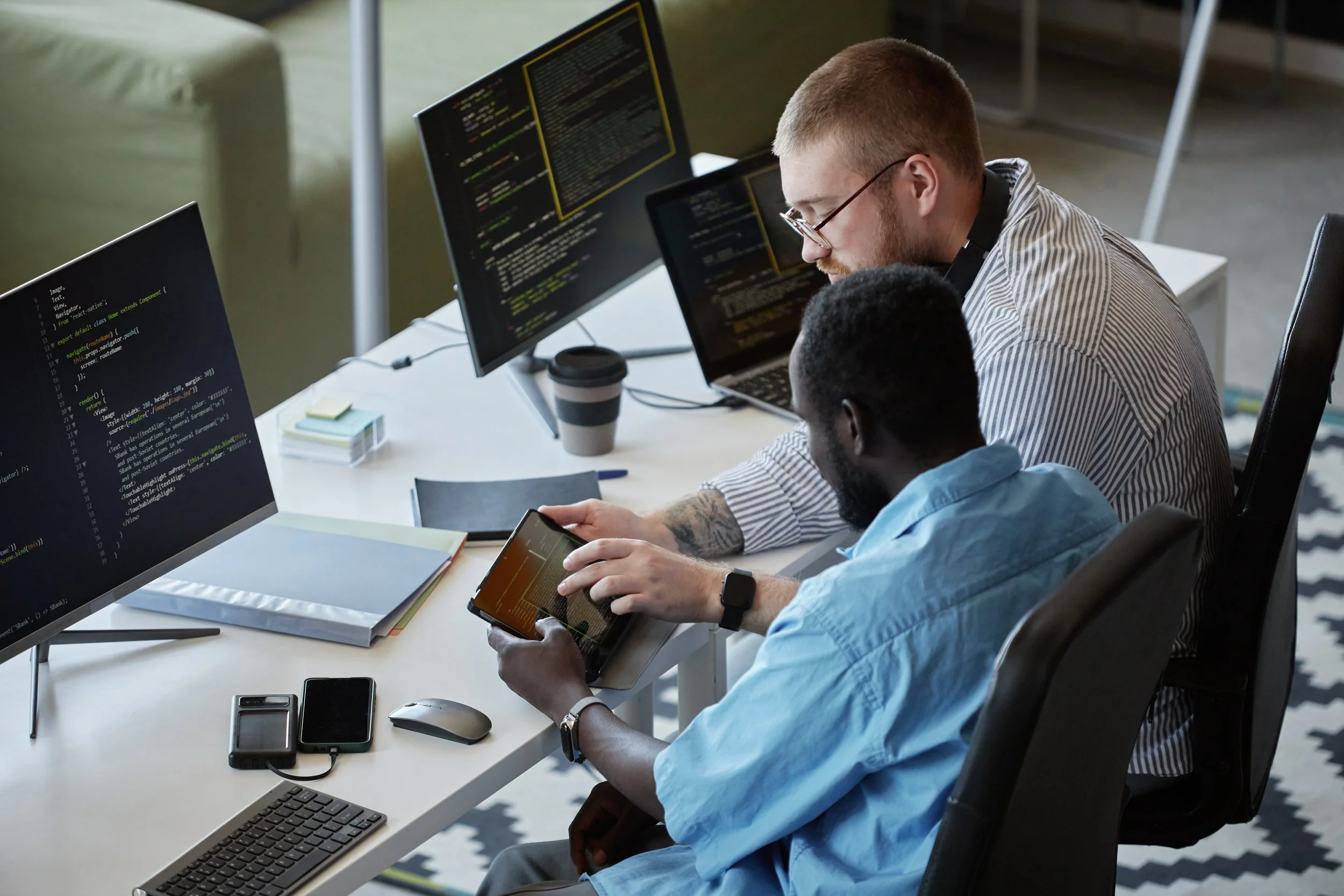 Two men working at a desk with multiple monitors, one using a tablet, the other using a smartphone, with coding displayed on screens.