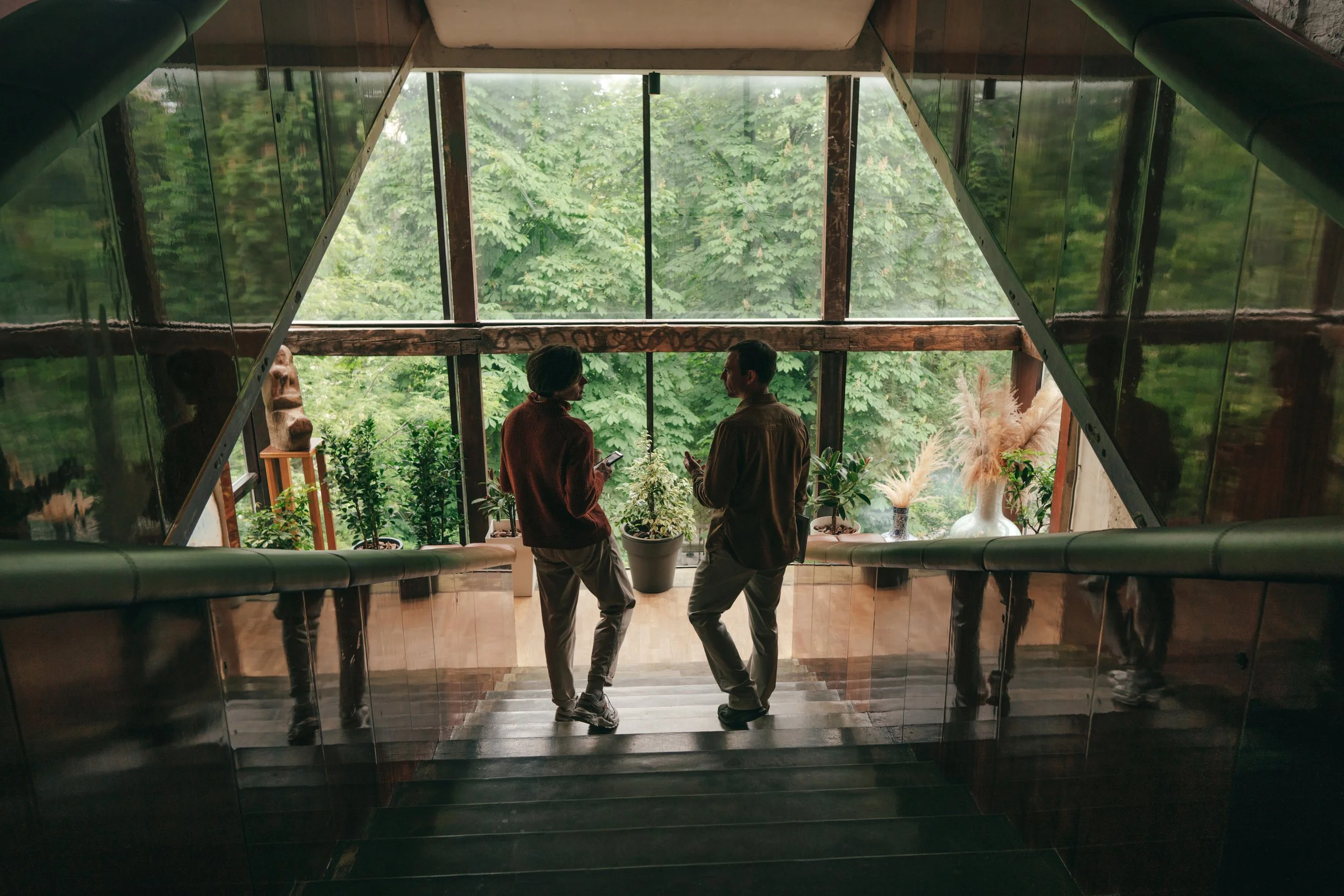 Two people stand on a staircase inside a building, facing large windows with green trees outside. They are engaged in conversation, one is holding a phone. The interior includes potted plants and decorative vases.