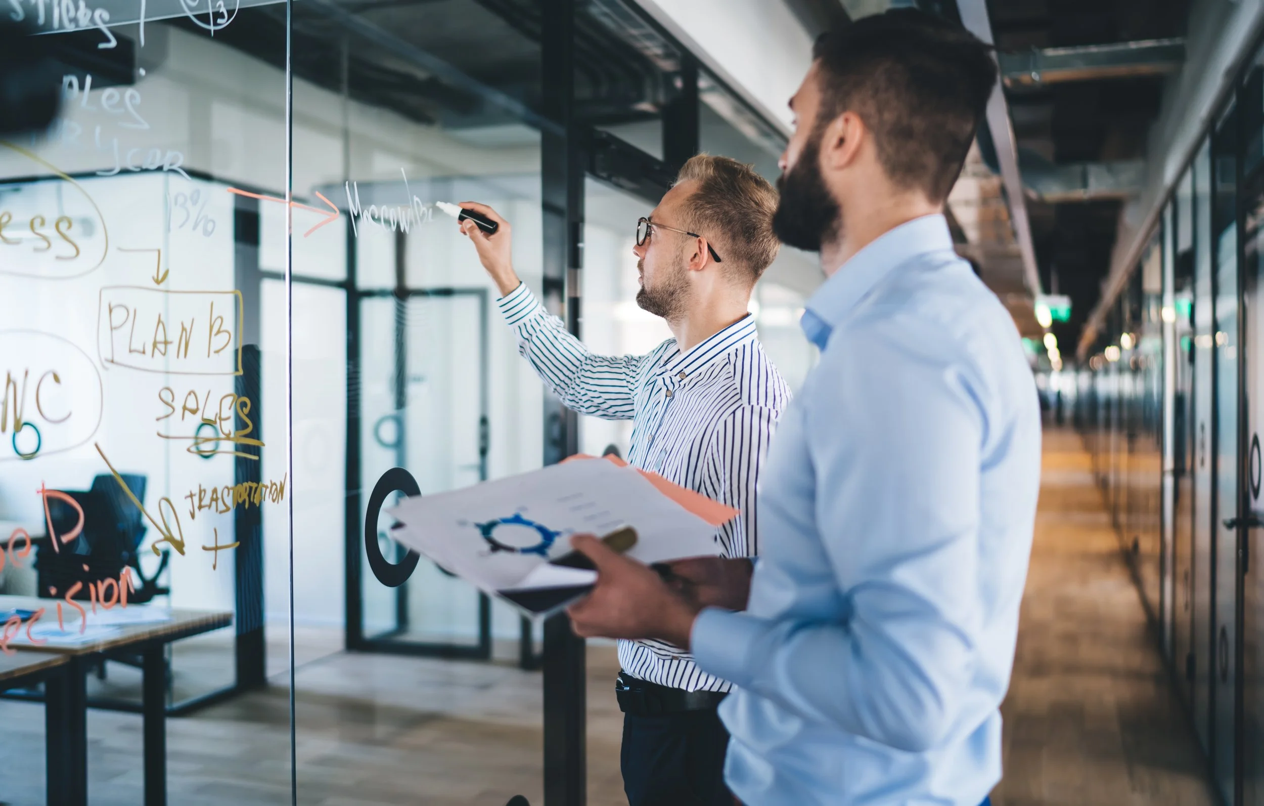 Two men in business attire working together in an office with glass walls, one is writing on a glass board while the other is holding a clipboard with papers.
