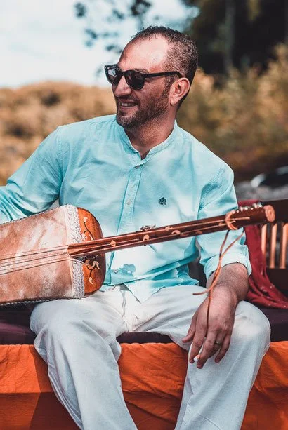 Black and white photo of a man with glasses and a beard, standing outdoors with arms crossed, looking to the side, with sunlight and shadow playing on his face.