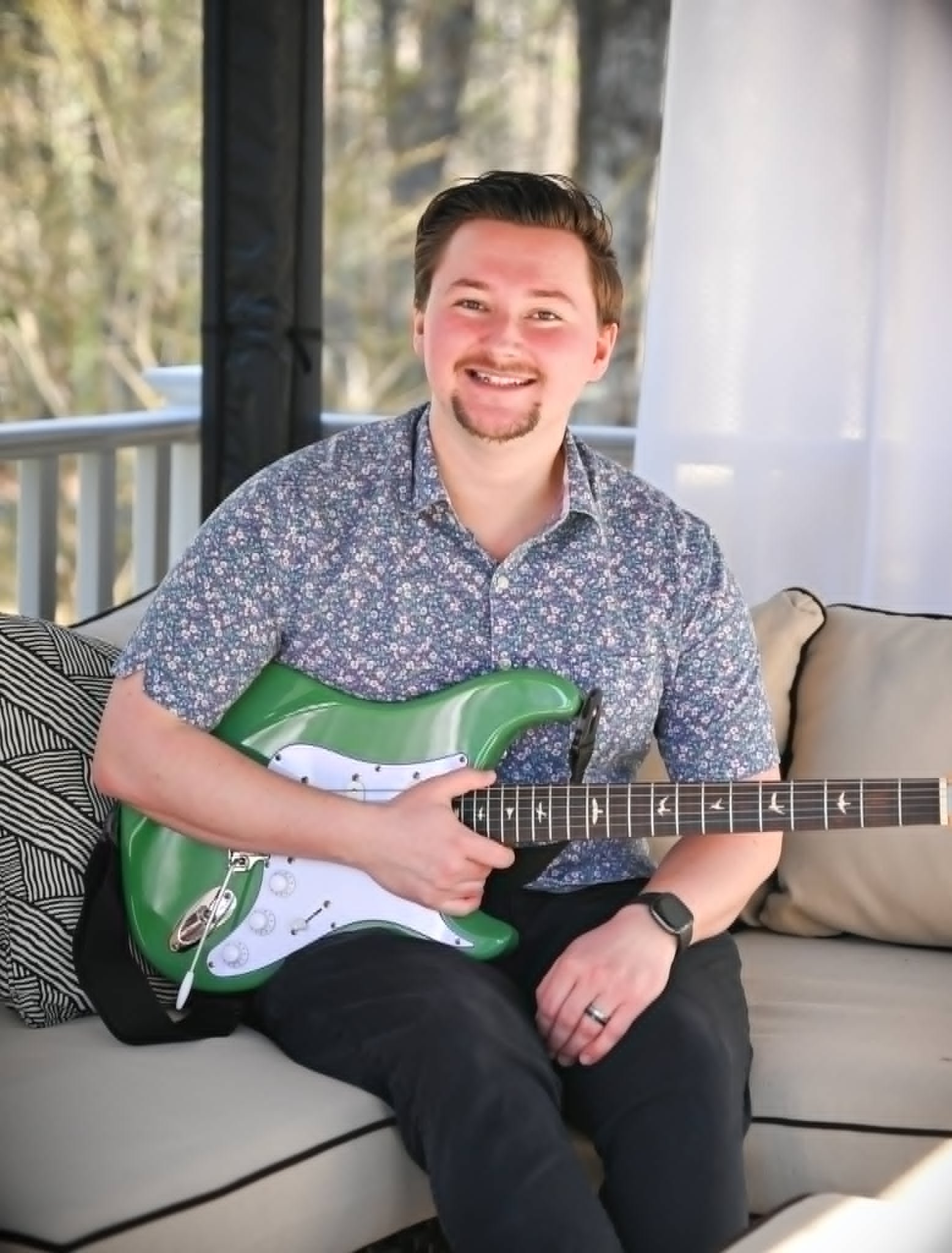 A man with brown hair, a goatee, wearing a patterned shirt, sitting on a beige couch with pillows, holding a green and white electric guitar, smiling at the camera, in a room with large windows and curtains.