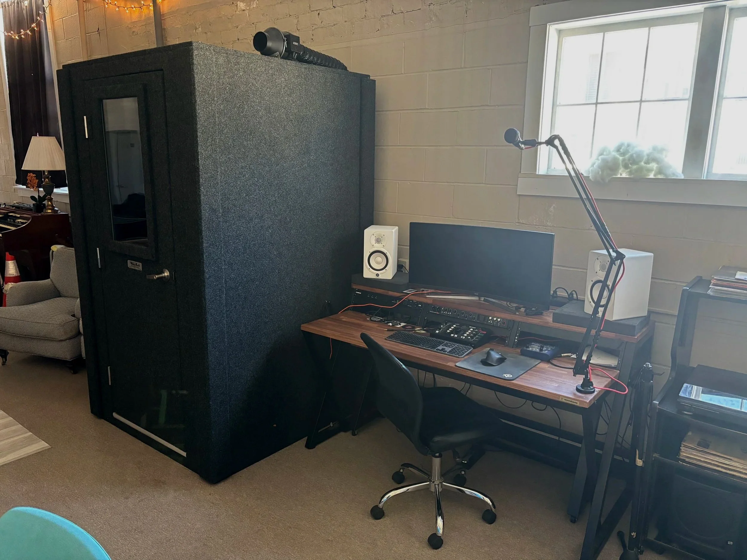 A room featuring a large black object, a microphone mounted on a desk, speakers, a computer monitor, a keyboard, a mouse, and audio equipment, with a window showing daylight and some clouds outside.