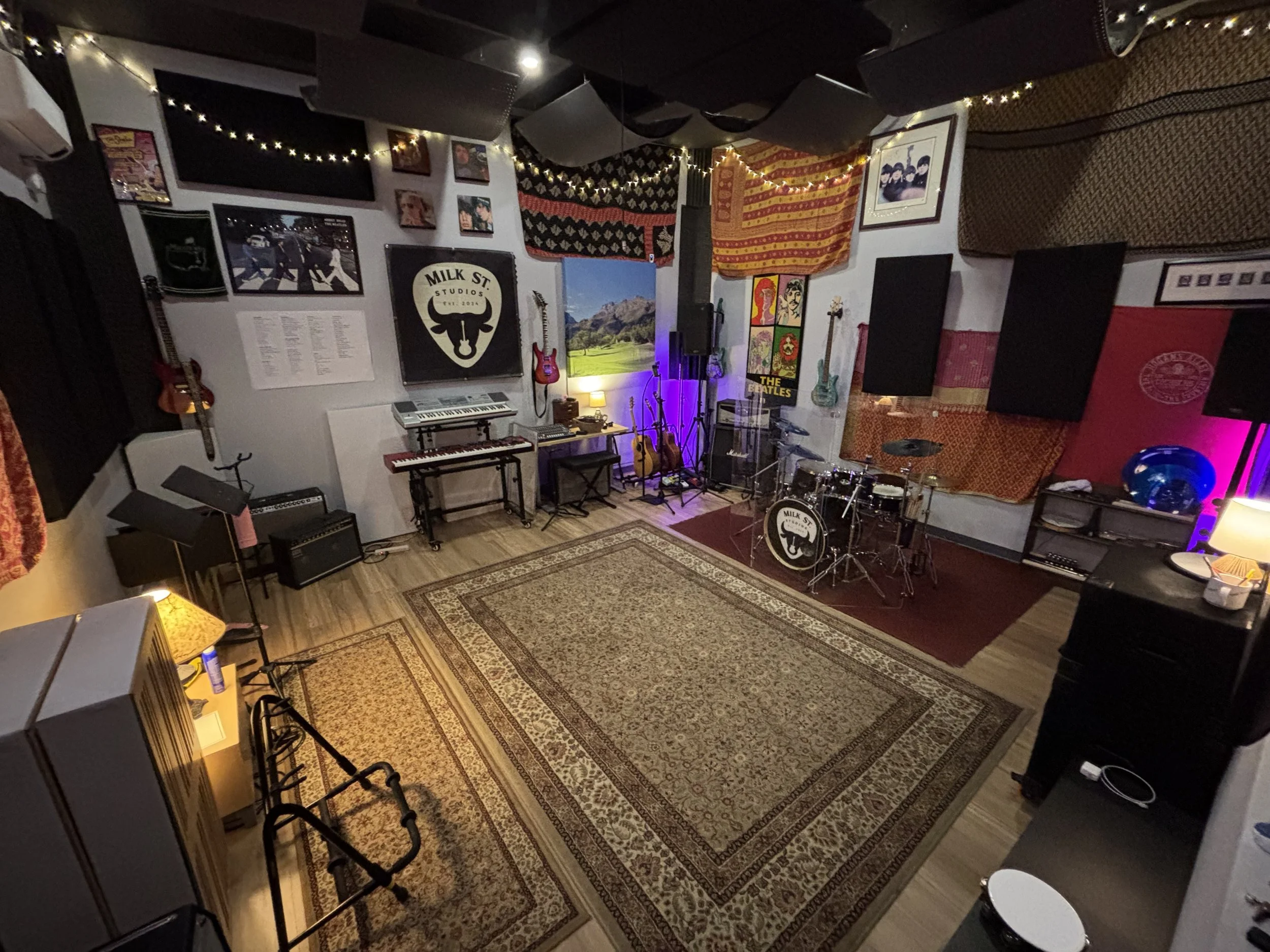 A music rehearsal or recording studio room with various musical instruments visible, including guitars on the wall, a keyboard, microphone, drums, and amplifiers. The walls are decorated with posters, artwork, and textiles, and the room has a cozy, artistic atmosphere with string lights and a patterned rug on the wooden floor.