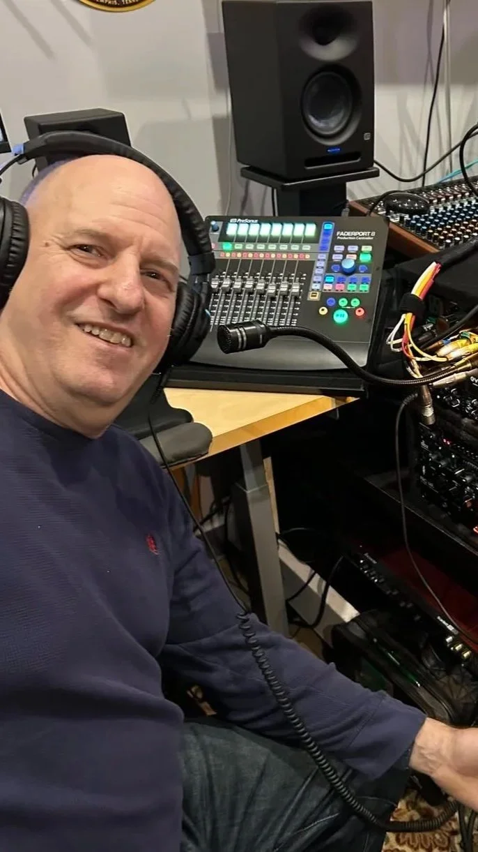 Man with headphones smiling in a recording studio with sound equipment, including a mixing console, audio rack, and speaker.