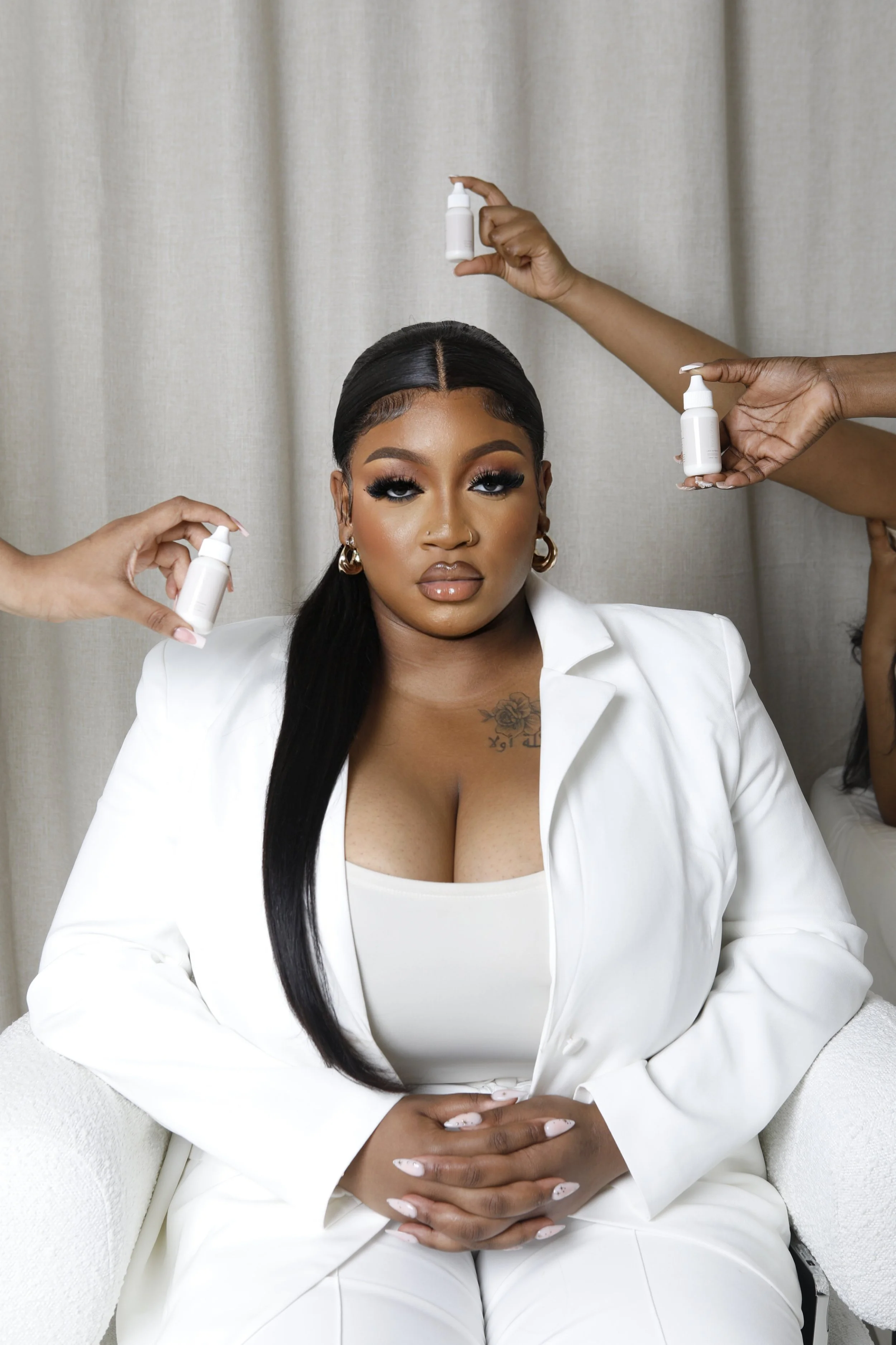 A woman with long black hair, wearing a white outfit, sitting on a white chair with her hands clasped on her lap, while three people around her hold small bottles of skincare or cosmetic products near her head and shoulders, in front of a neutral beige curtain.