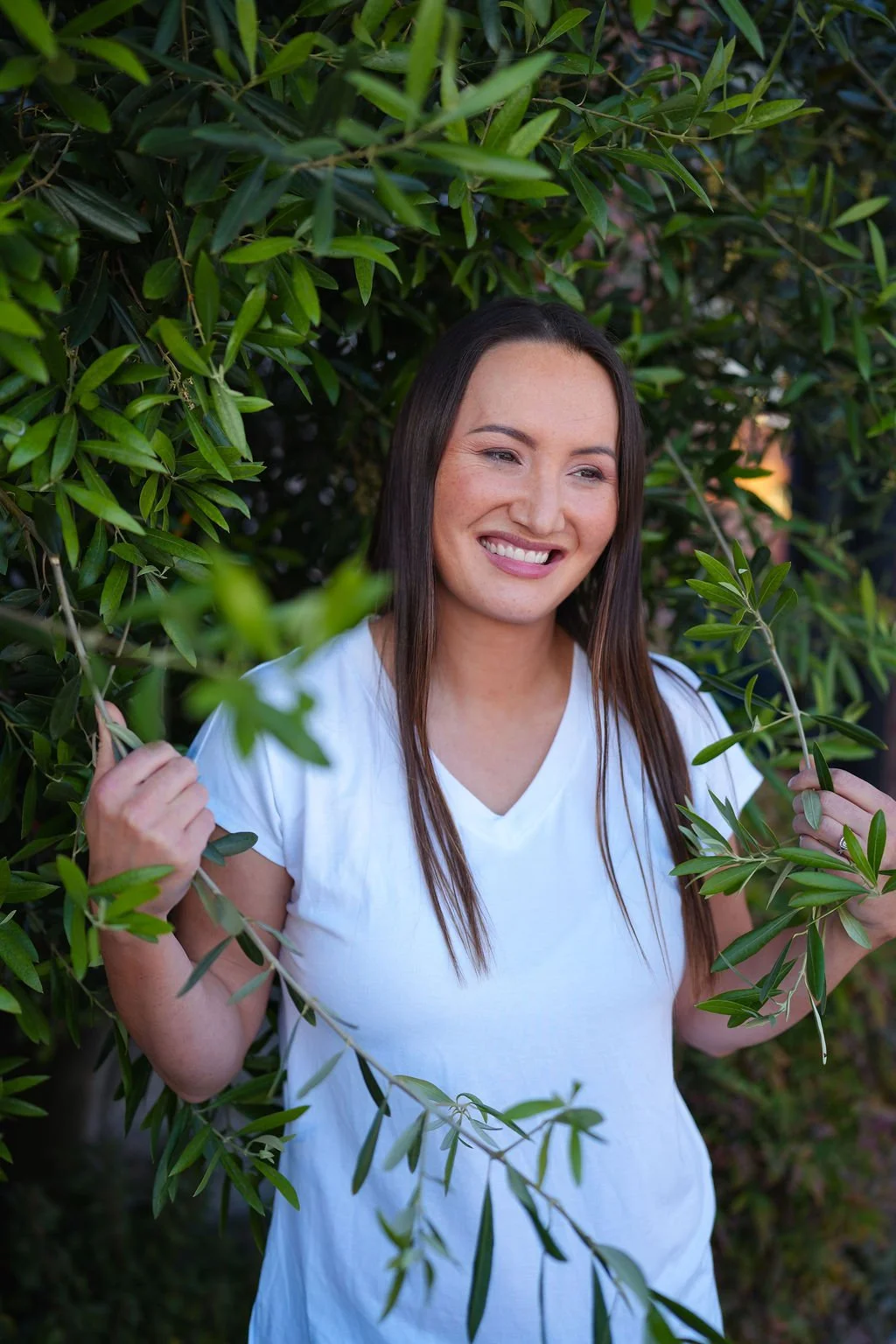 A woman with long brown hair and a white t-shirt standing amid green leafy foliage, smiling and holding branches of the plant.