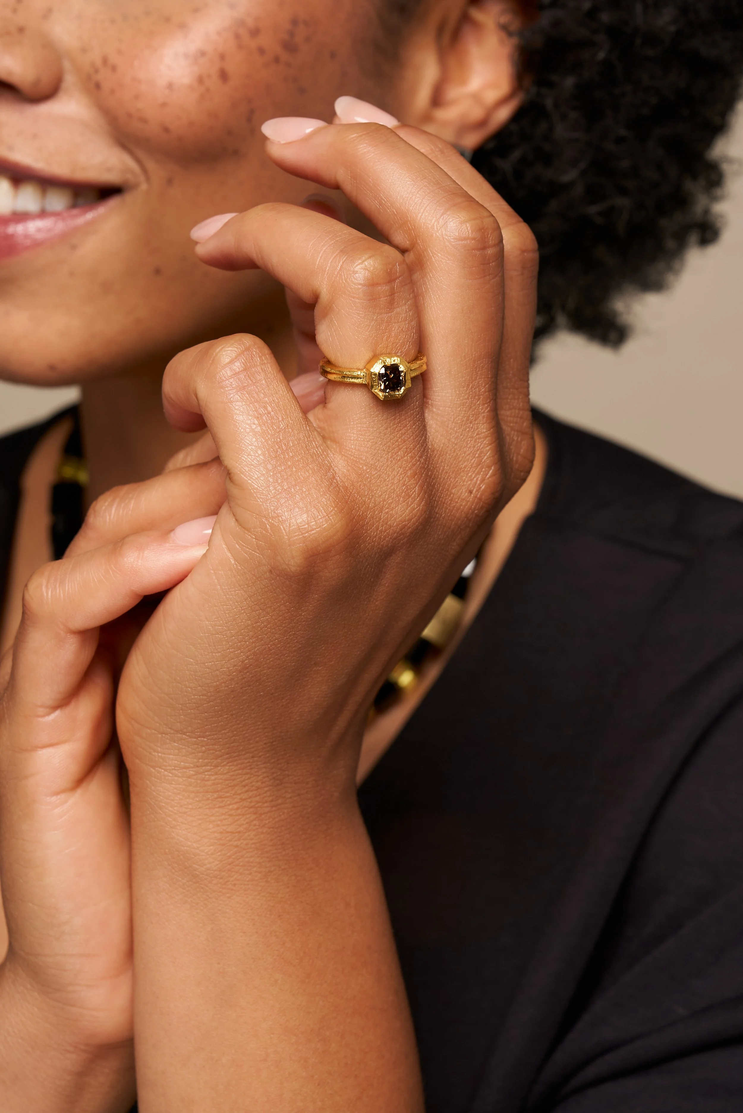 Close-up of woman's hand with a gold ring featuring a black gemstone, near her face showing freckles, smile, and part of her black outfit and jewelry.
