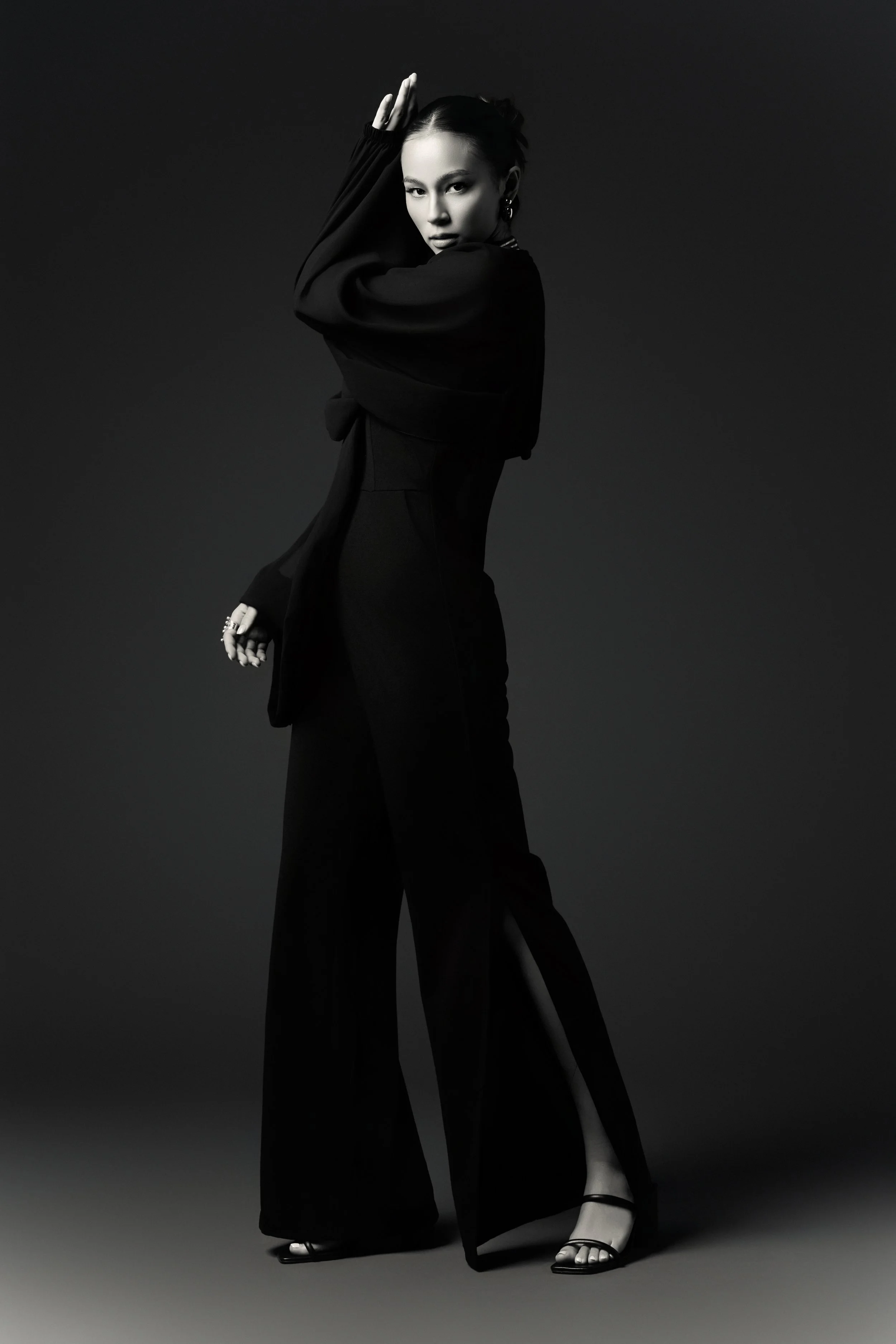 A woman dressed in black posing with one hand on her head, looking intensely at the camera, in a black-and-white studio photograph.