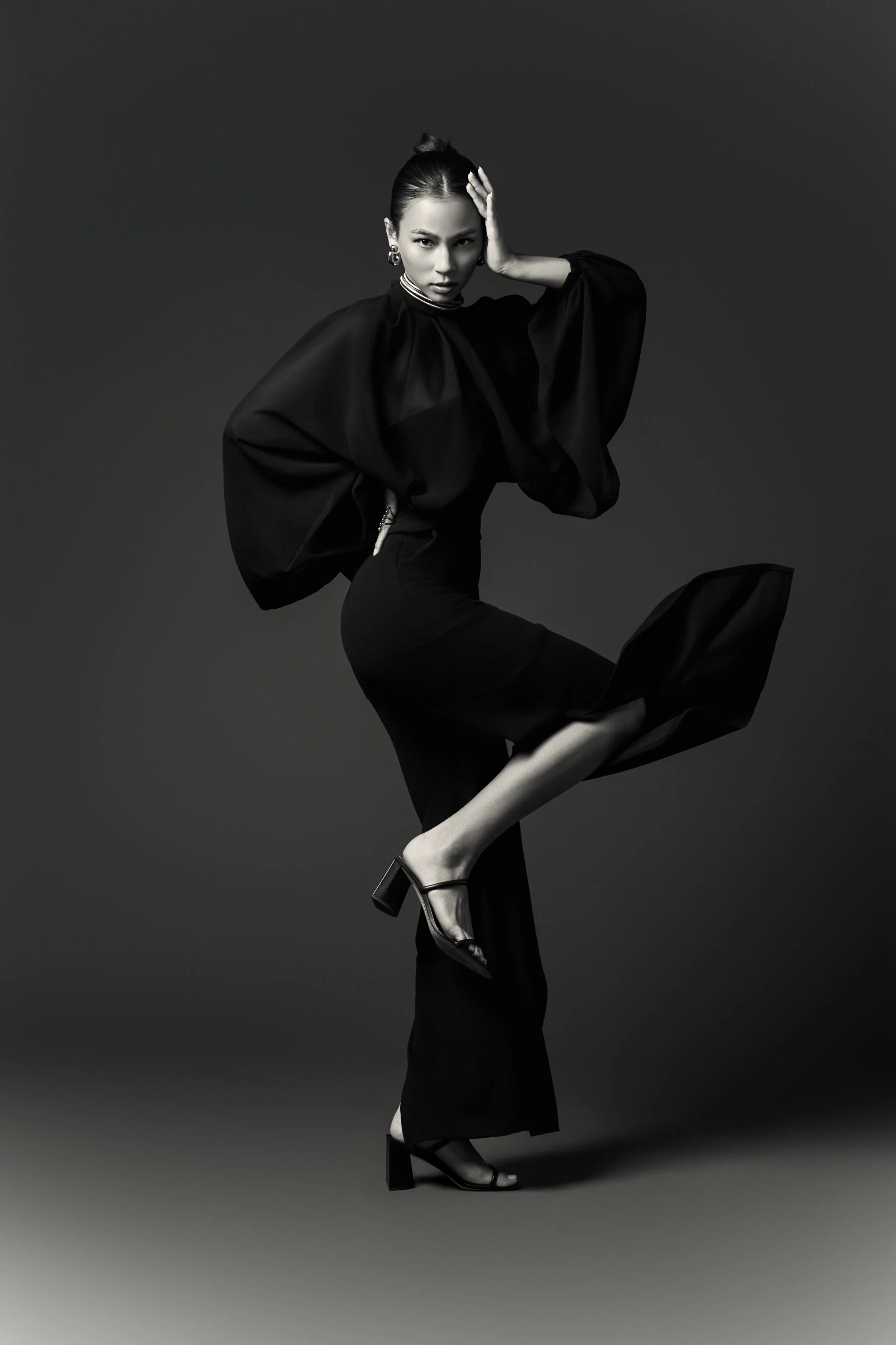 A woman in a black dress and high heels striking a pose on a plain background.