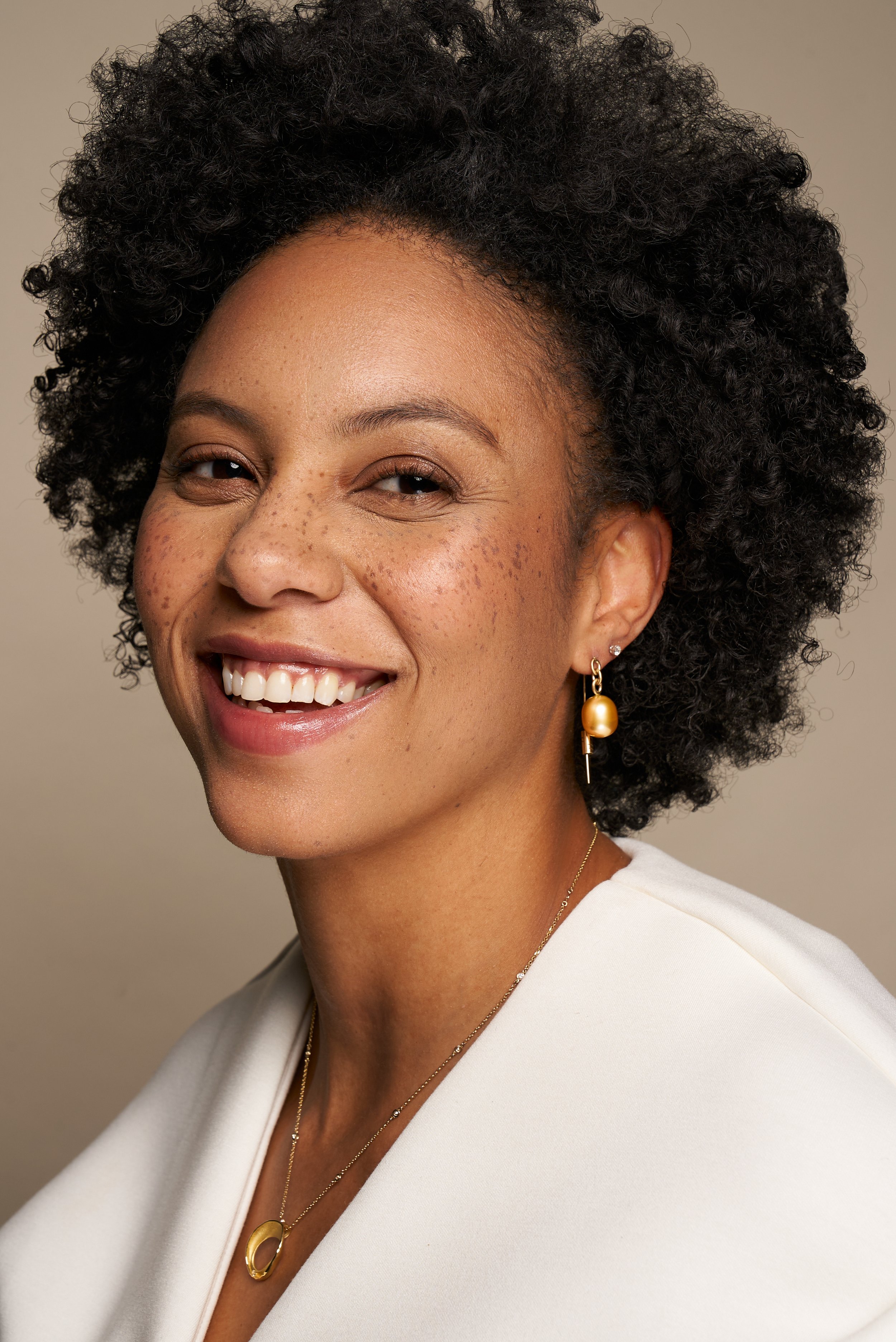 Close-up portrait of a smiling woman with natural curly hair, gold earrings, and a gold necklace, wearing a white blazer.