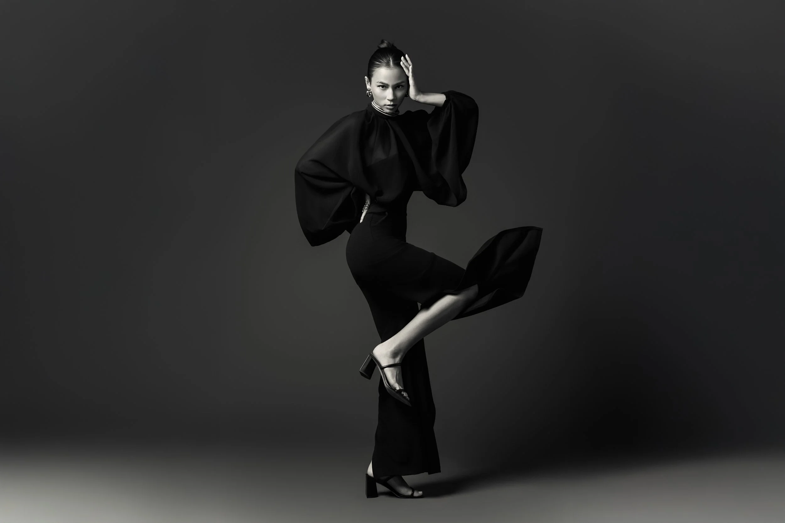 A woman dressed in black, striking a high-knee pose on one leg, with her hand on her head and looking at the camera, against a dark background.