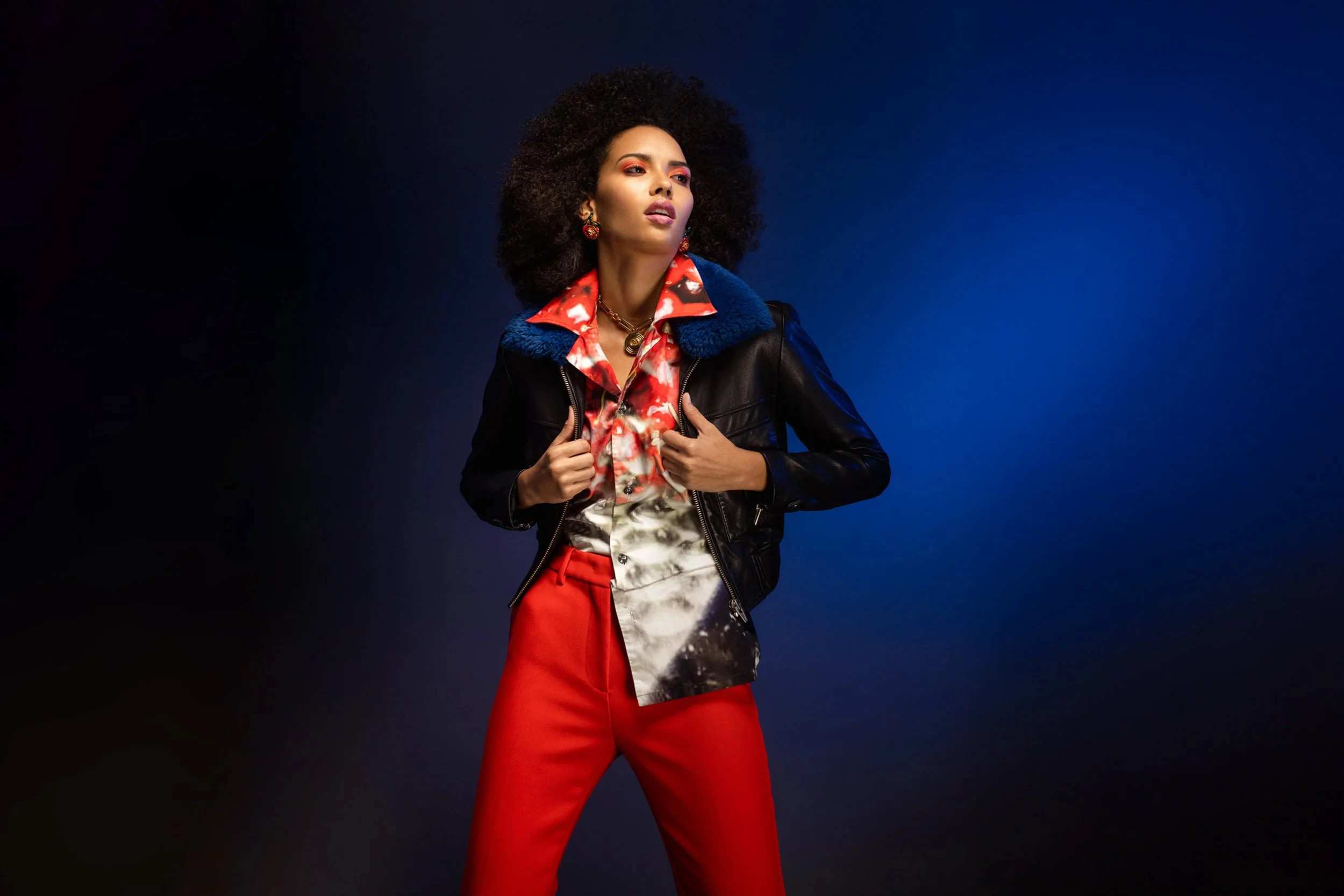 Fashion model with voluminous curly hair wearing a colorful patterned shirt, black leather jacket with blue collar, and red pants against a dark blue background.