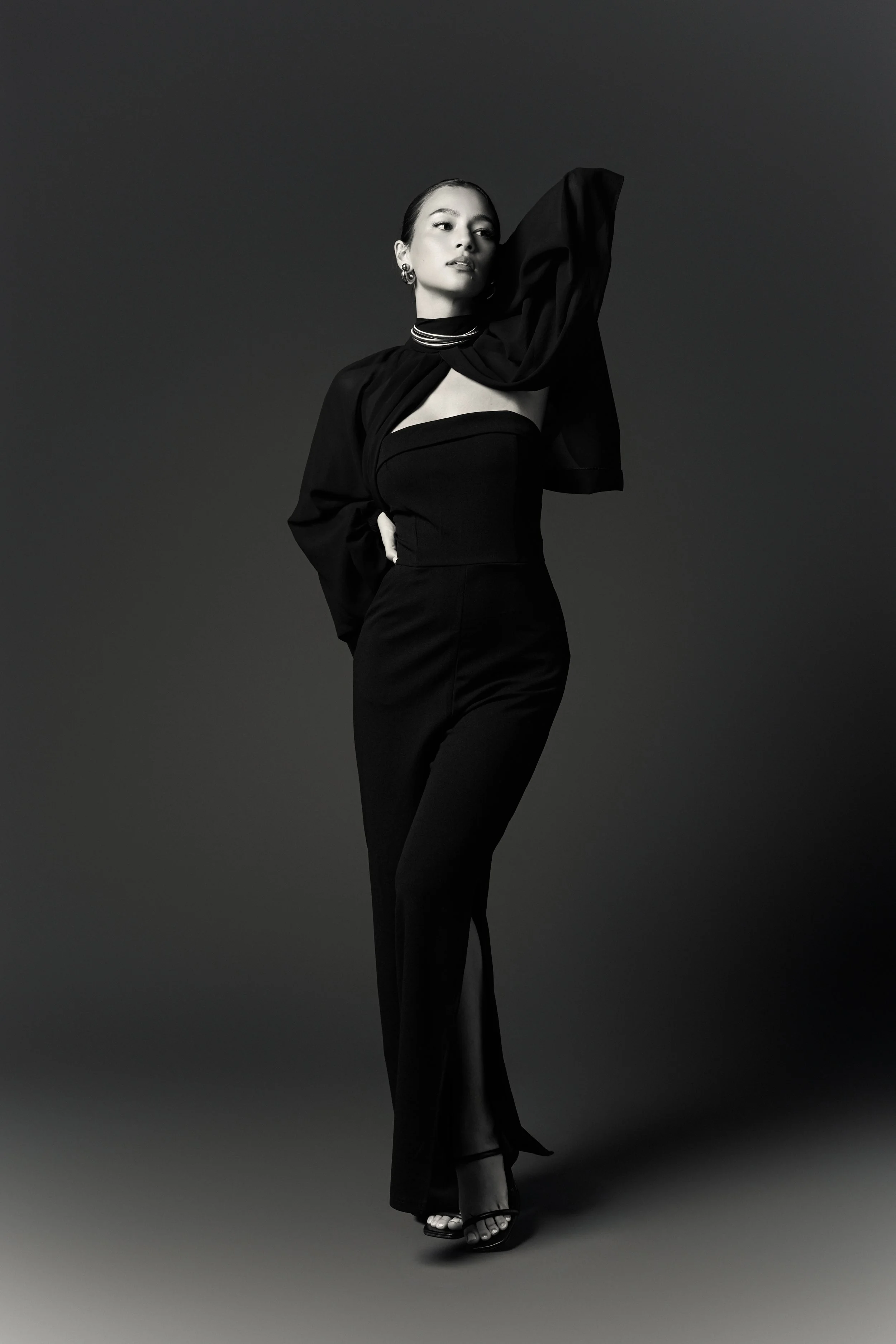 A woman in a black strapless jumpsuit posing with her right arm raised and her left hand on her hip, wearing high heels and earrings, looking confident in a studio with a dark background. Alonza Mitchell