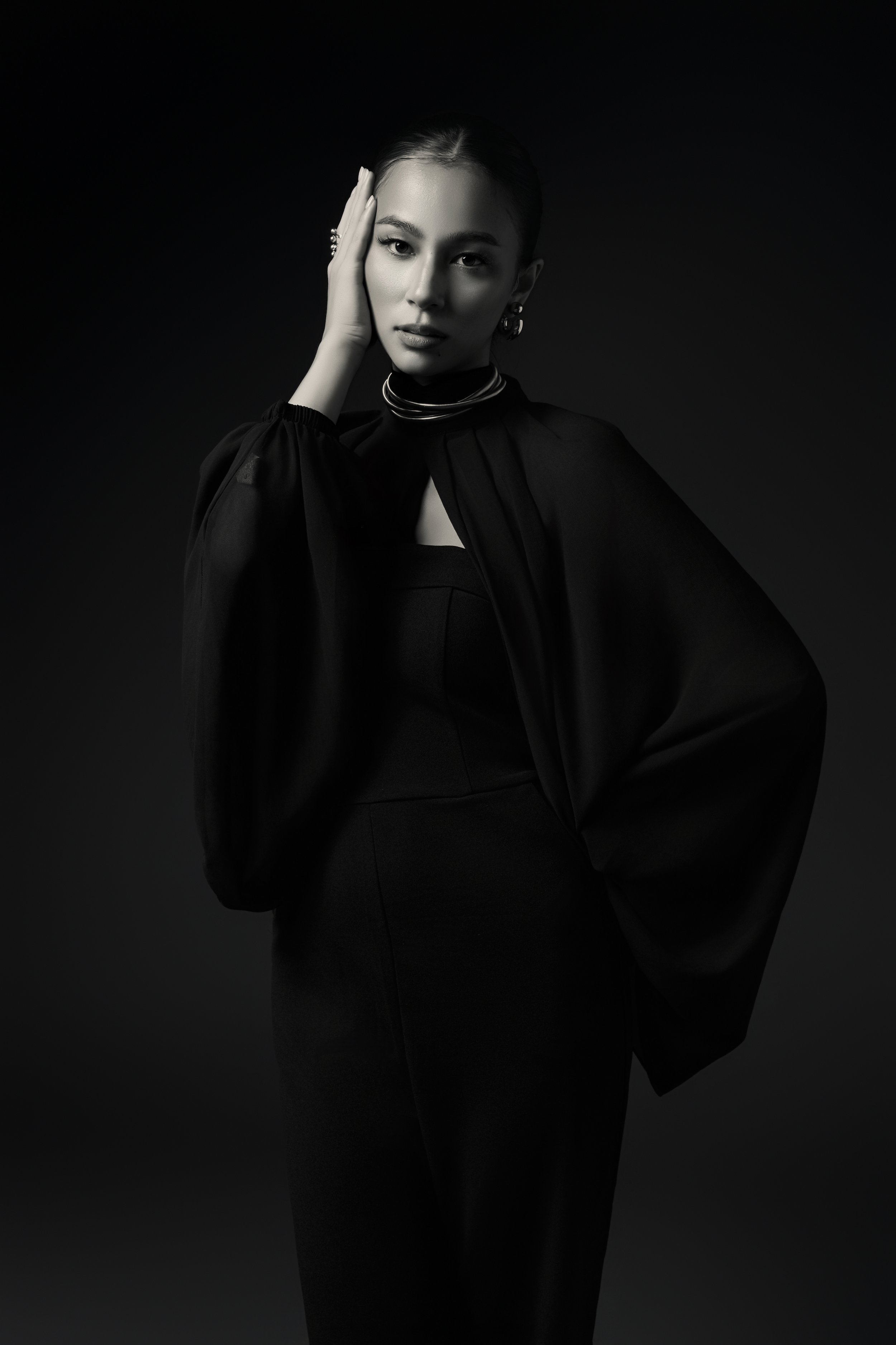 A black and white portrait of a woman with styled hair, wearing jewelry and elegant clothing, touching her face with her left hand.