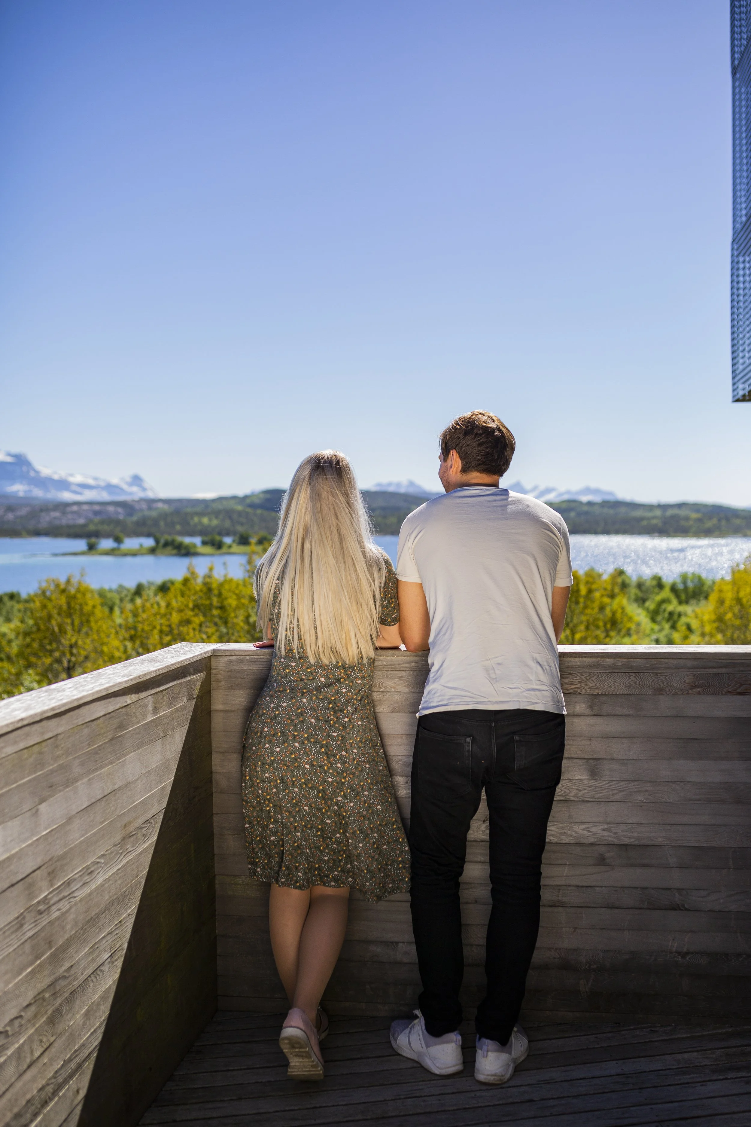 To personer står på en treplatting og nyter utsikten over innsjø, fjell og blå himmel på en solrik dag.