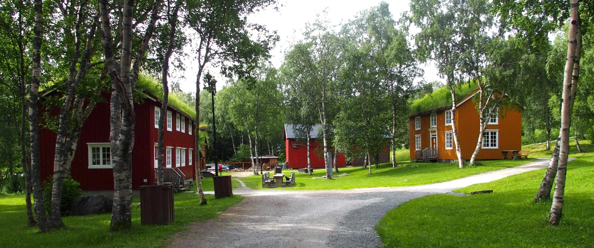 Fargerike hus omgitt av grønne trær og en grusbane, med benker og søppelkasser i et avslappende utendørsmiljø.
