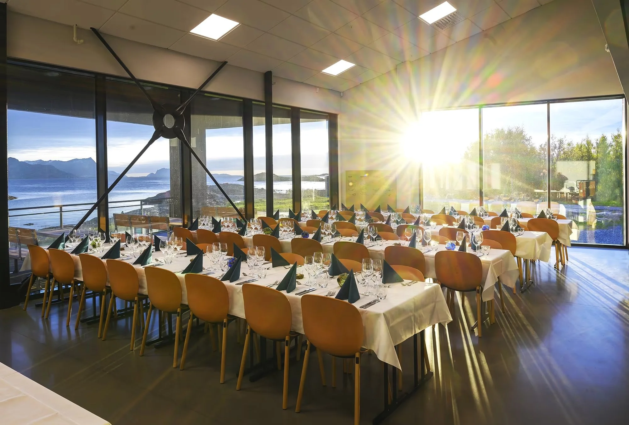 Innendørs konferanserom med langbord dekket til middag, store vinduer med utsikt over sjø og fjell, solnedgang eller soloppgang, spiseutstyr og pyntet til arrangement.