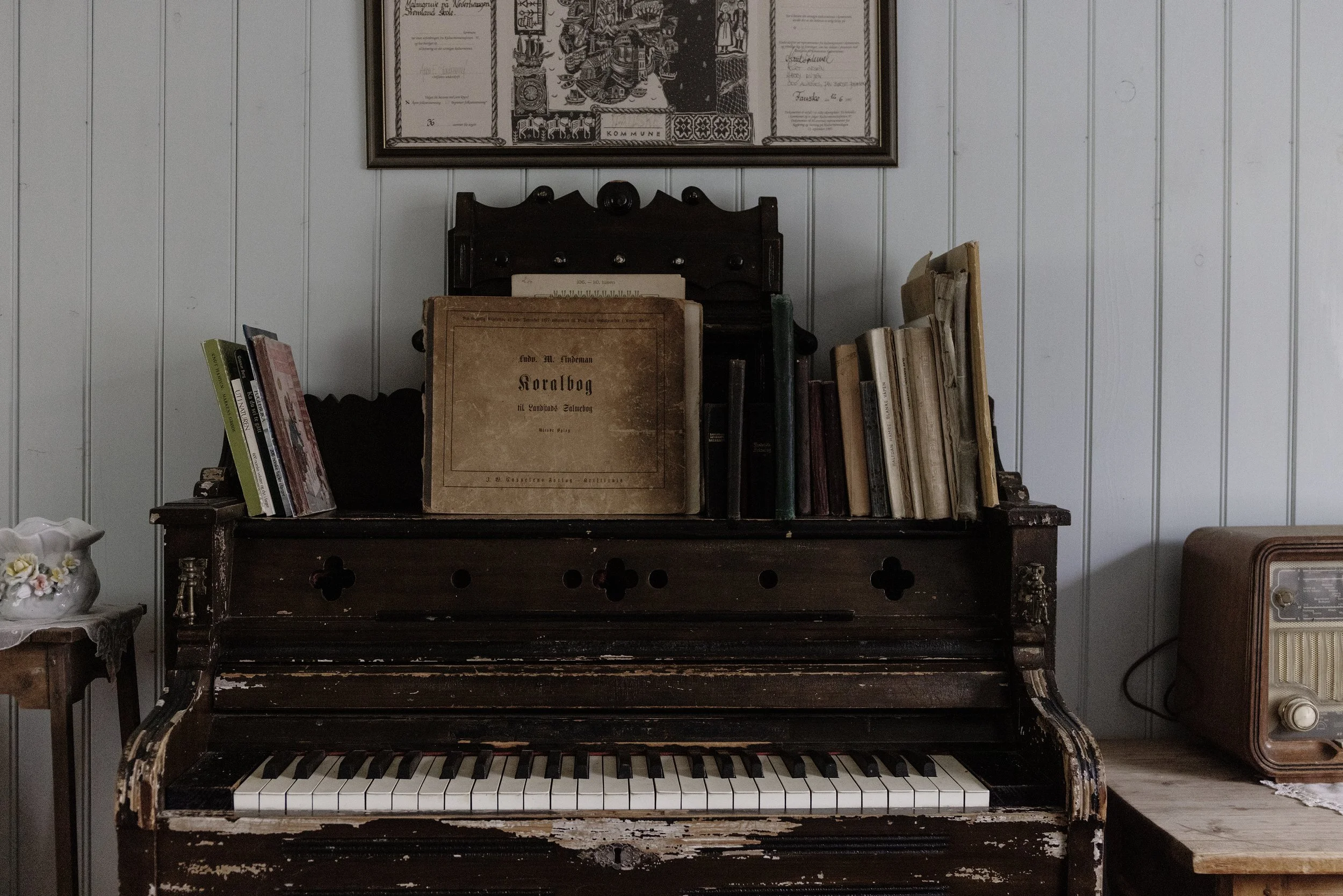 Ein altes, abgenutztes Klavier mit einigen Büchern darauf steht an einer Wand mit vertikalen Paneelen. Über dem Klavier hängt ein gerahmtes Bild oder Dokument.