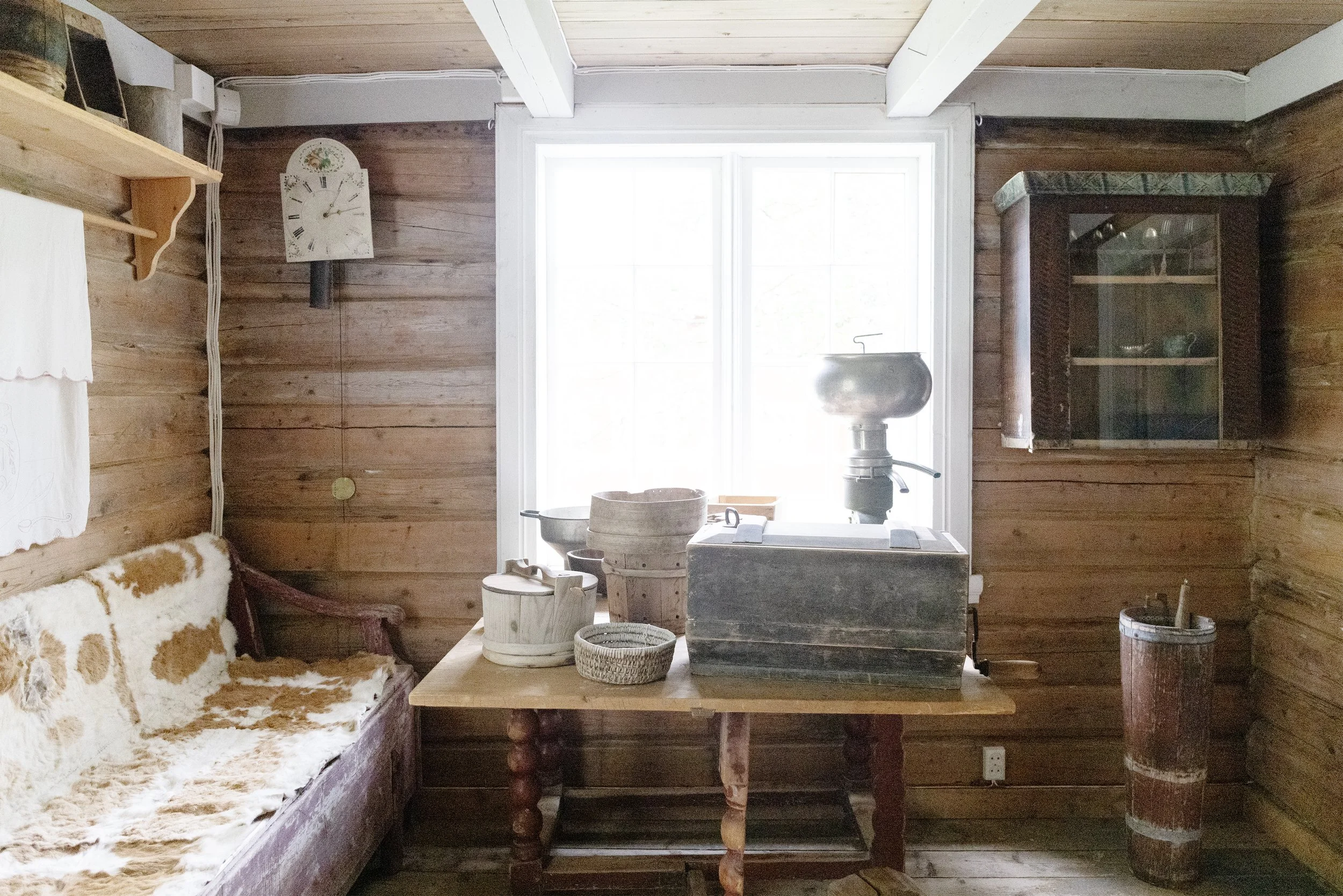 Interior of a room with wooden walls, window and various old objects such as a water pump, baskets, and a cupboard.