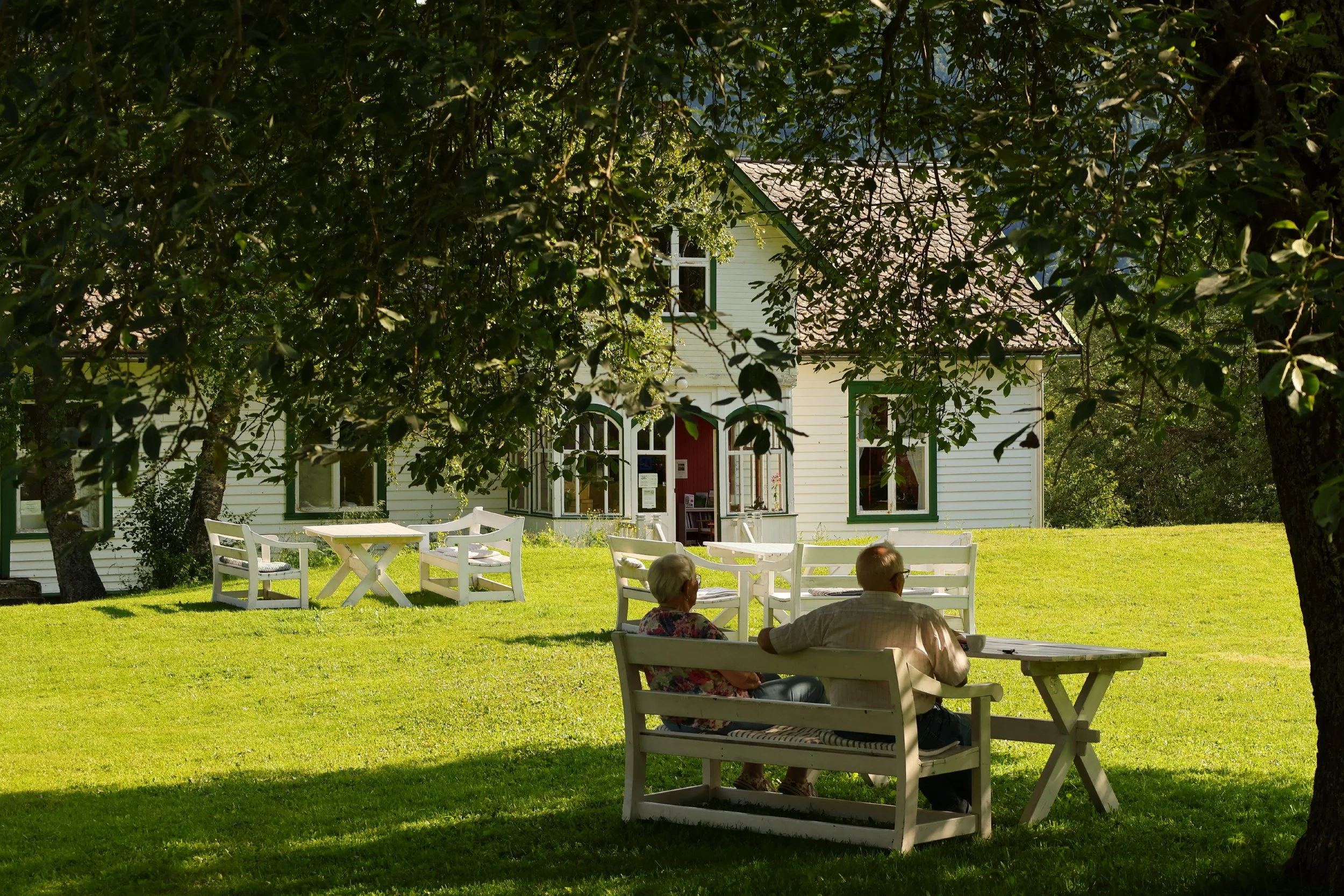 To eldre personer som sitter på benker i en hage, omgitt av grønne trær, foran et hvitt hus med grønne vindusrammer.