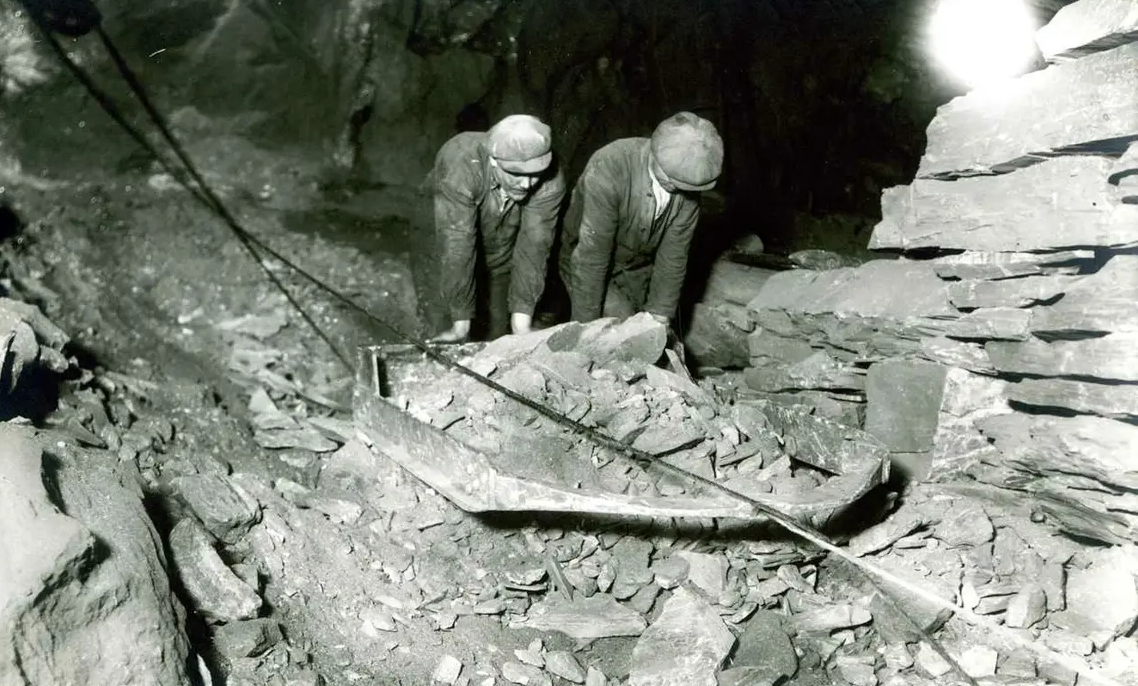 Two traditional miners wearing helmets and work clothes, standing by a cart filled with rock in a mountain cave or mine in the 19th century.