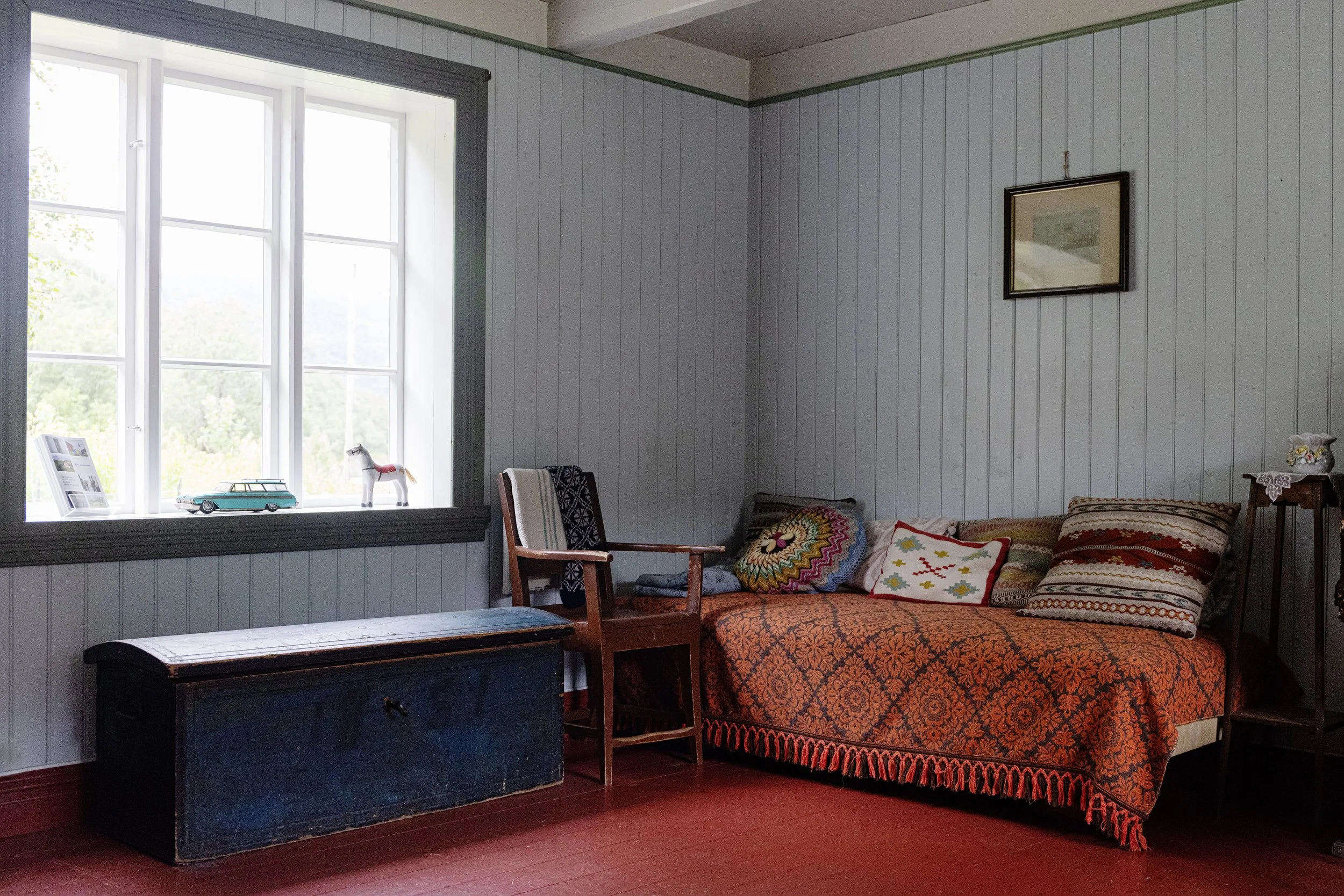 Ein Zimmer mit Fenster, mehreren Stühlen, einem Bett mit verschiedenen Kissen und einem alten Koffer auf dem Boden.