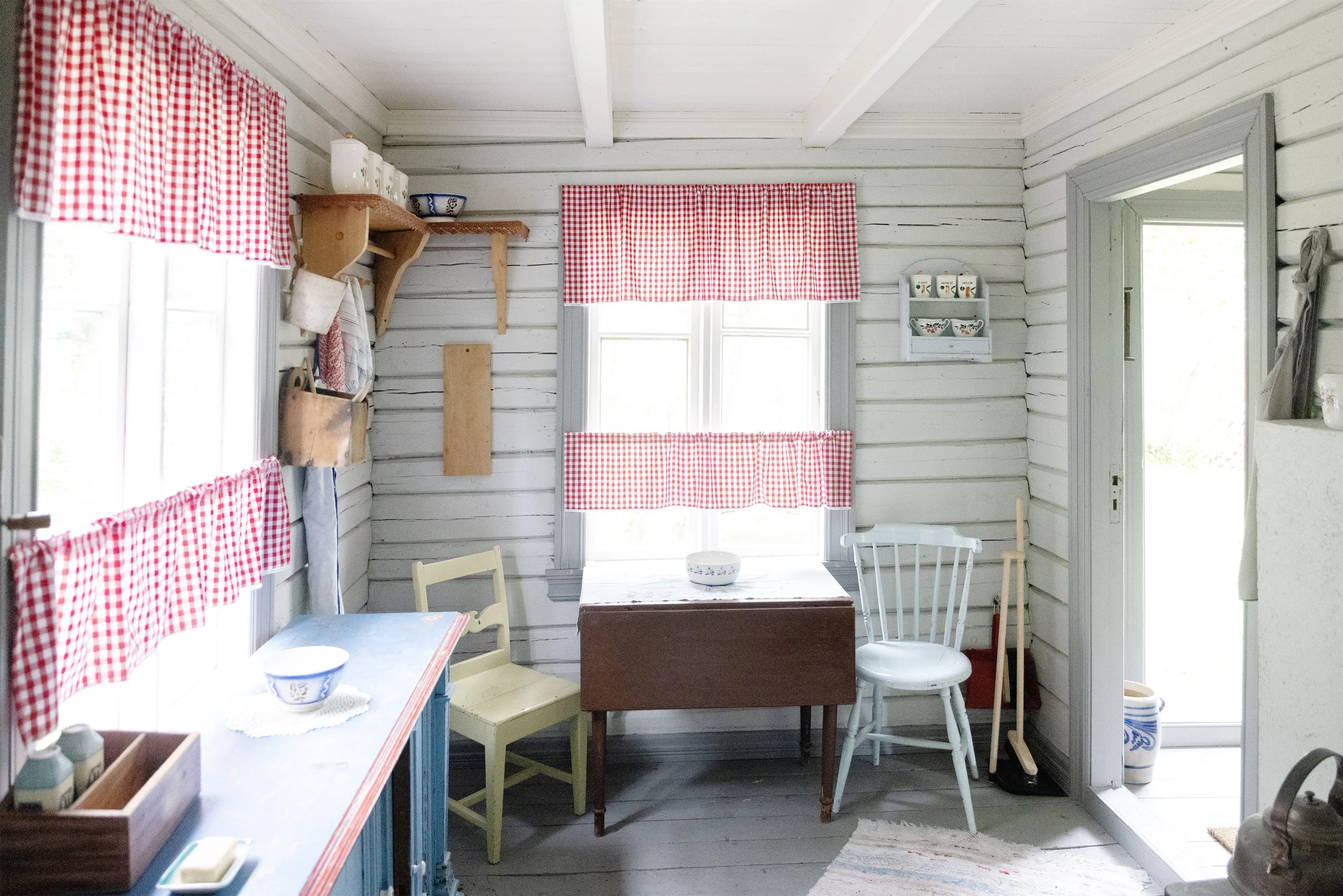A bright and cozy kitchen with white wood-paneled walls, red-checkered curtains, and small furniture, including a table, chairs, and a shelf of cups.