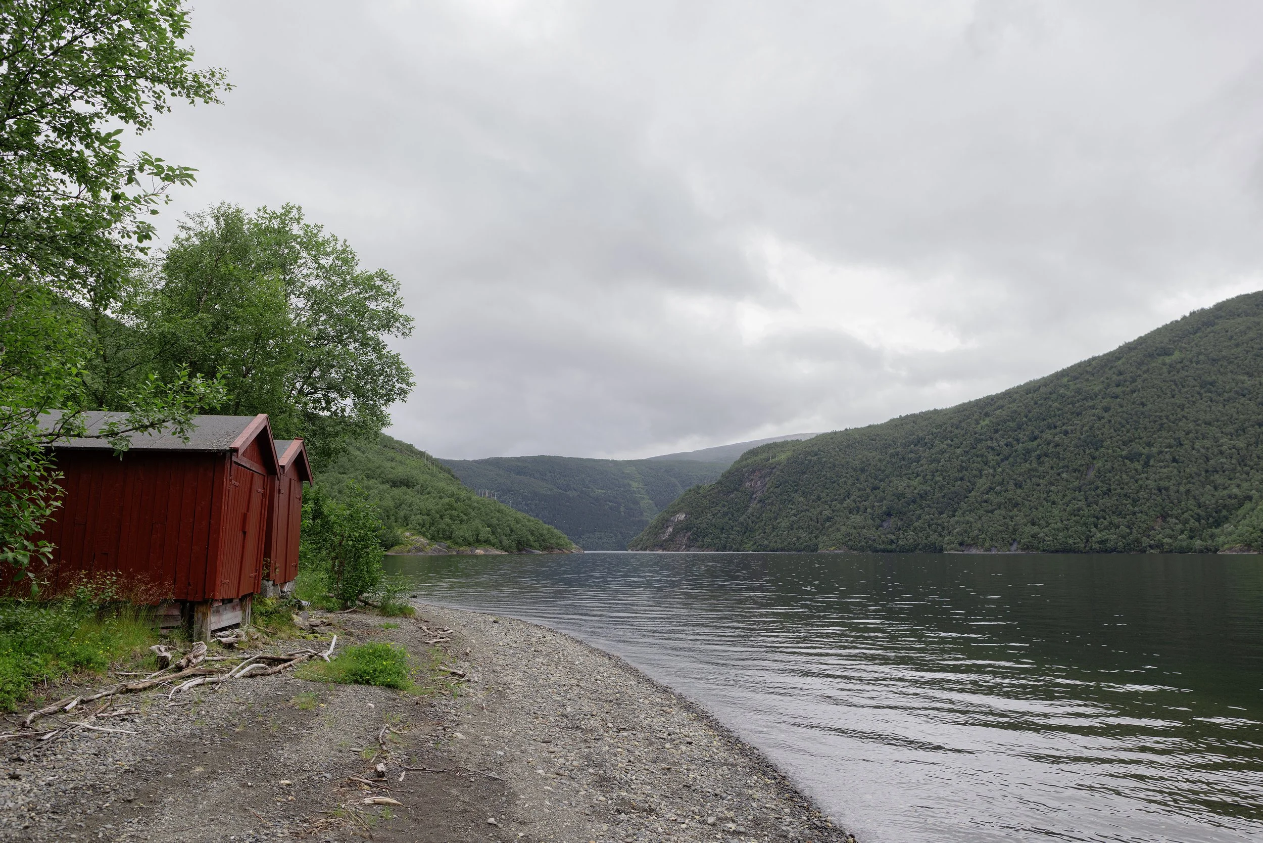 Eine versteckte, kleine rote Hütte am Wasser, umgeben von grünen Bäumen, mit Bergen im Hintergrund unter einem bewölkten Himmel.