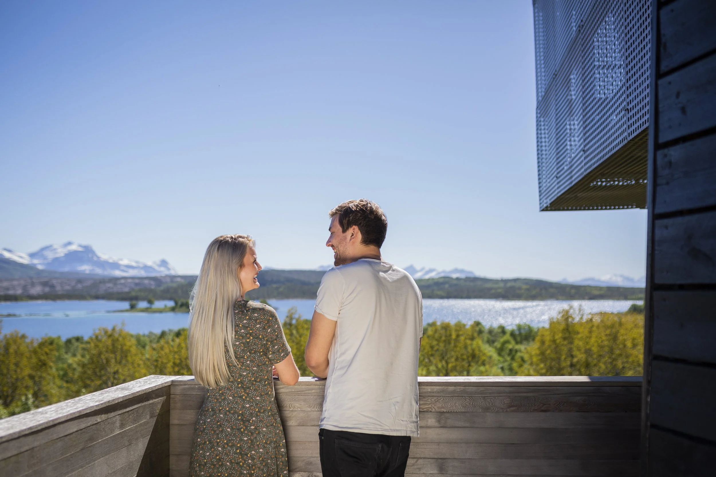 Modern, geometric building overlooking lake, surrounded by green forests and mountains in the background under a blue sky with clouds.