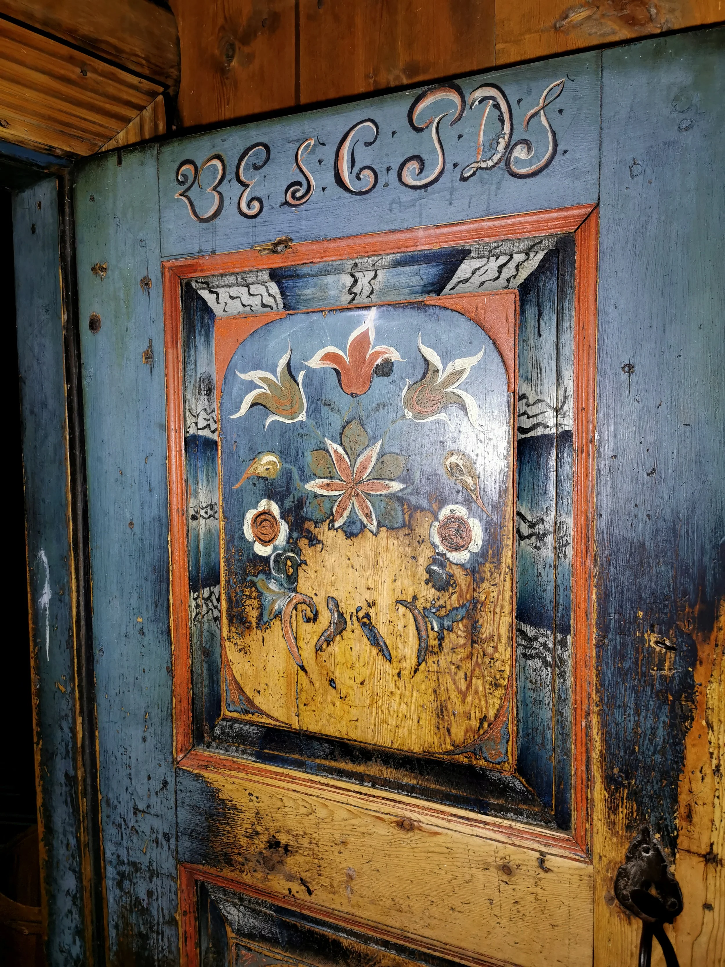 A vintage grey and blue floral wallpaper, with a large, old-fashioned, red and gold painted wall clock on the left, and old black and white photographs of people in gold frames on the wall.