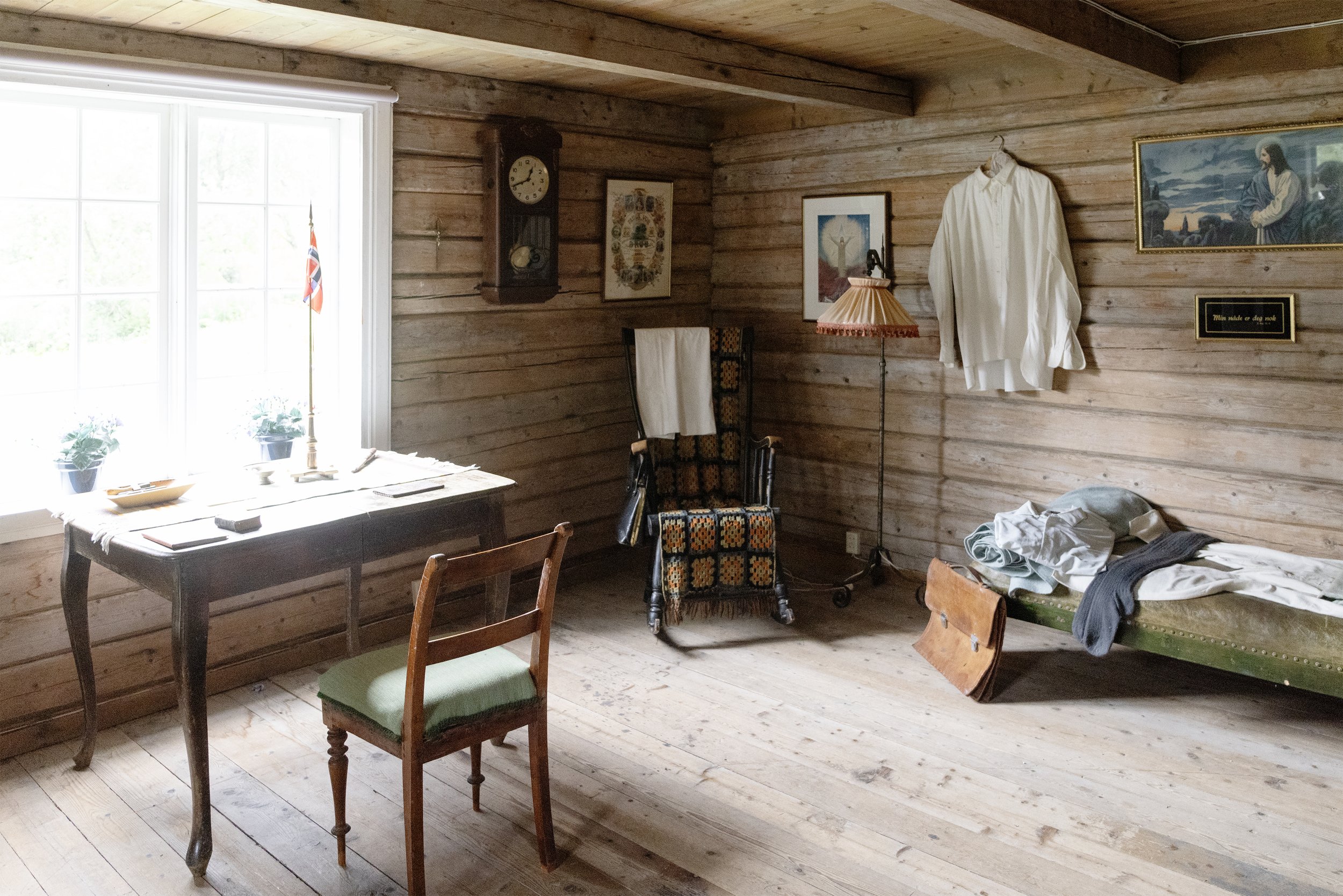 Interior of a room with wooden walls, a table with textiles and a chair, pictures and decorations on the wall, a clothesline hanging, and a room next to it with a bed and clothes.
