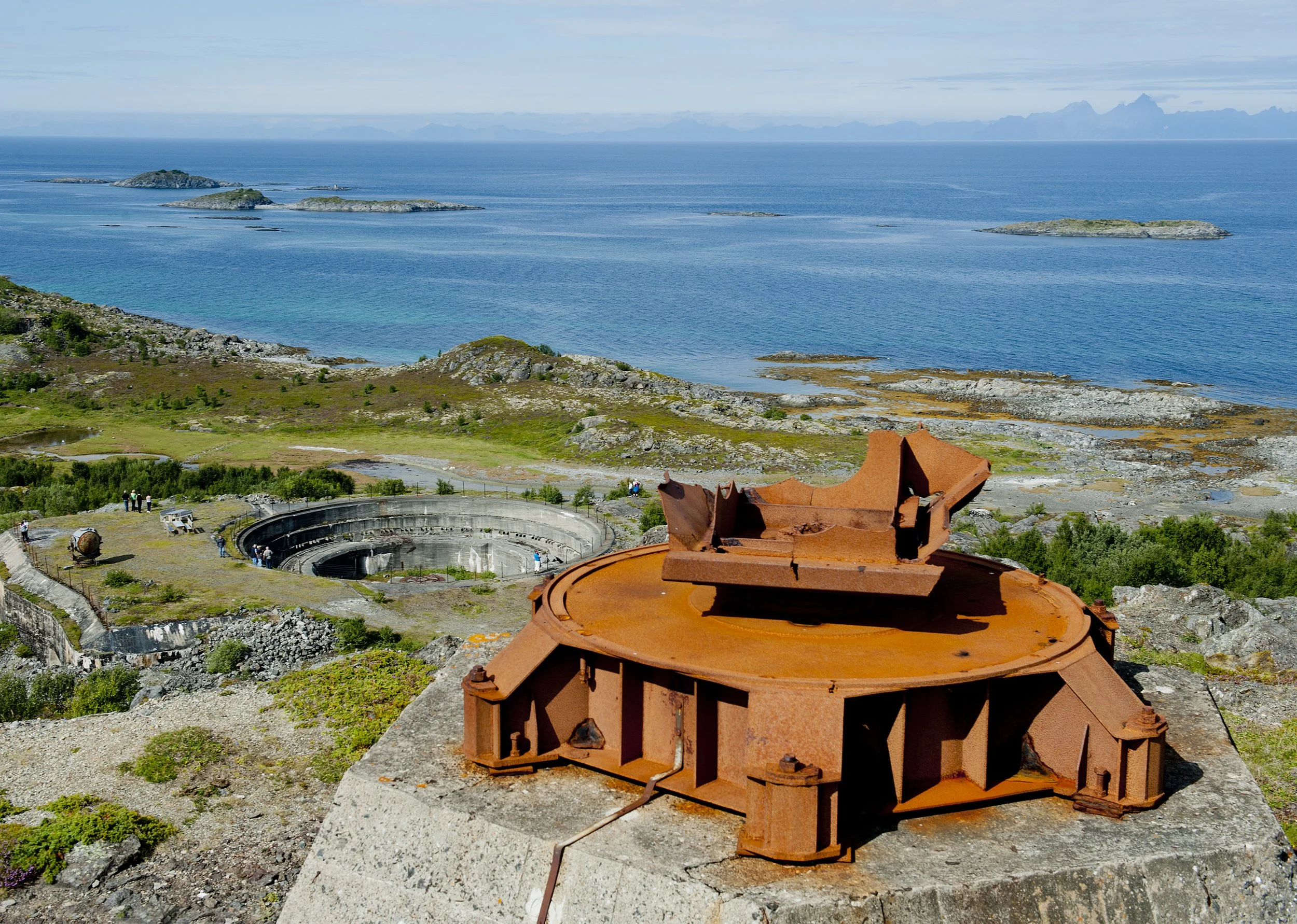 Rusty, old military turret station on a rocky hilltop overlooking an ocean with islands and a circular bunker below.