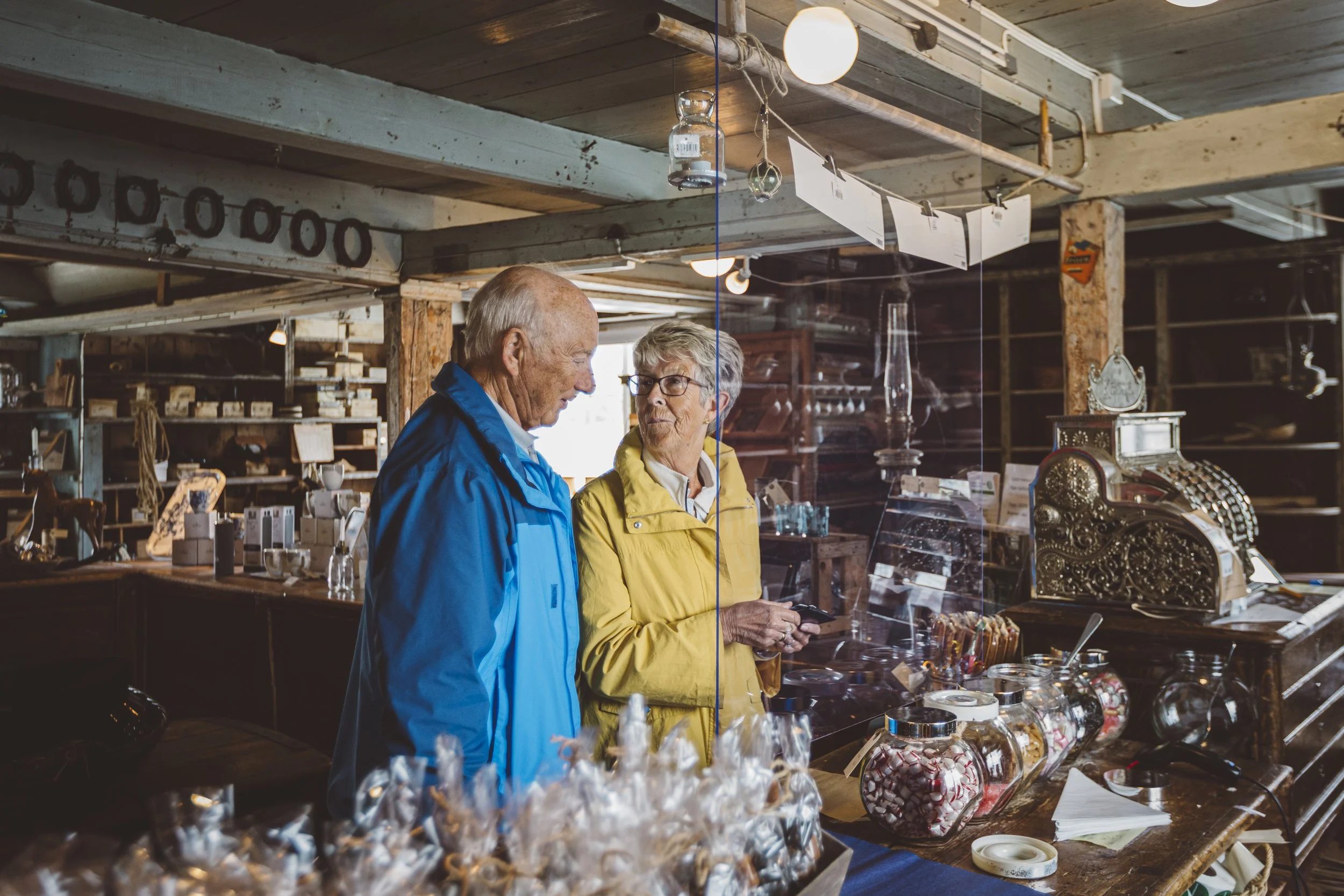 To eldre personer, en mann i blå jakke og en kvinne i gul jakke, står i en butikk med gamle objekter og godteri. De ser på hverandre bak en glassvegg.