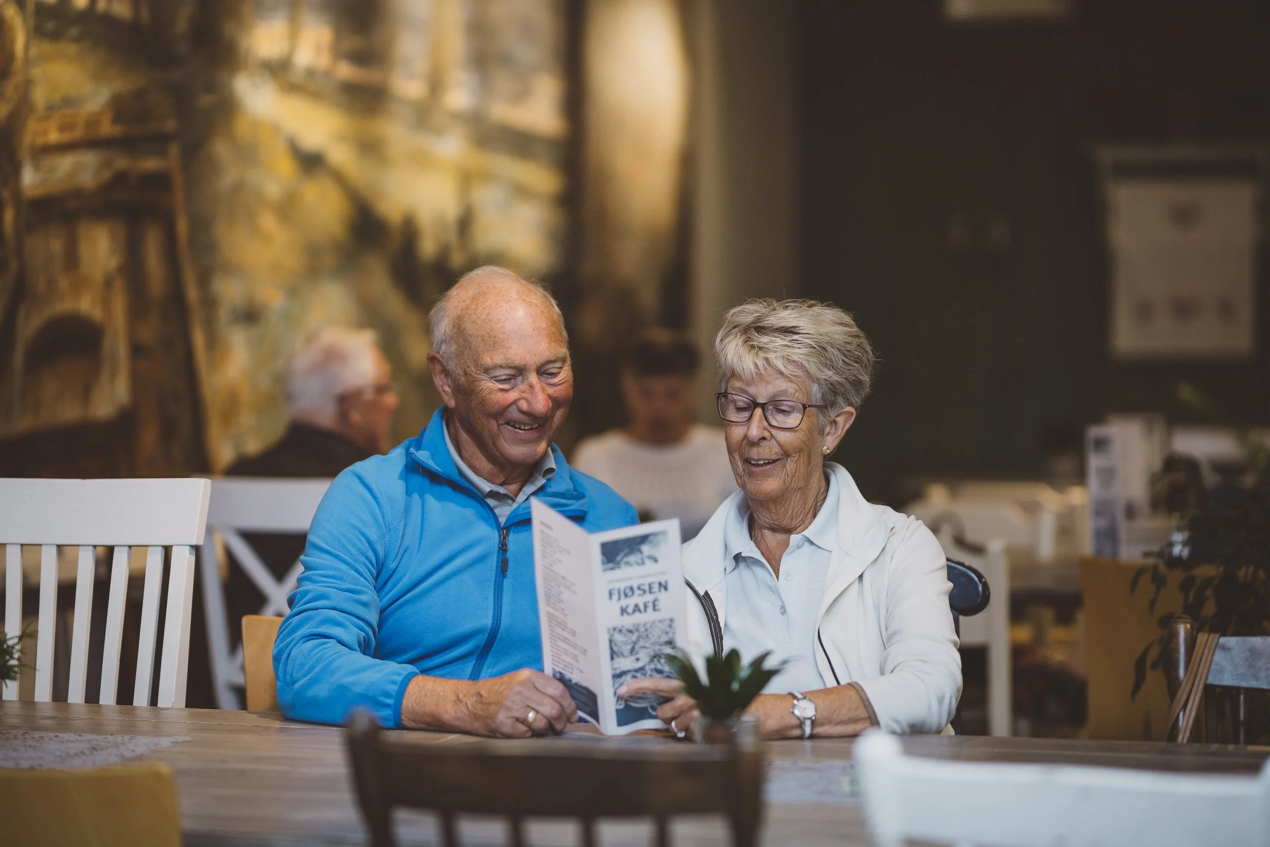 Zwei ältere Menschen, ein Mann und eine Frau, lächeln und lesen gemeinsam in einem Café eine Broschüre.