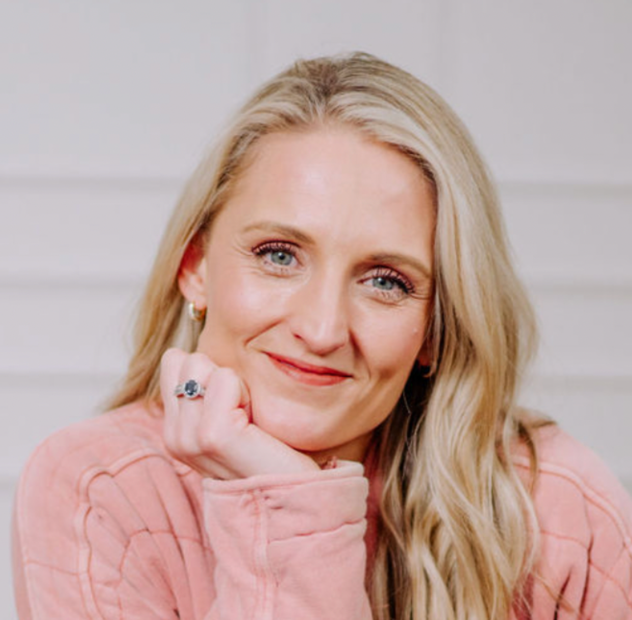Smiling woman with long blonde hair wearing a pink sweater, resting her chin on her hand.