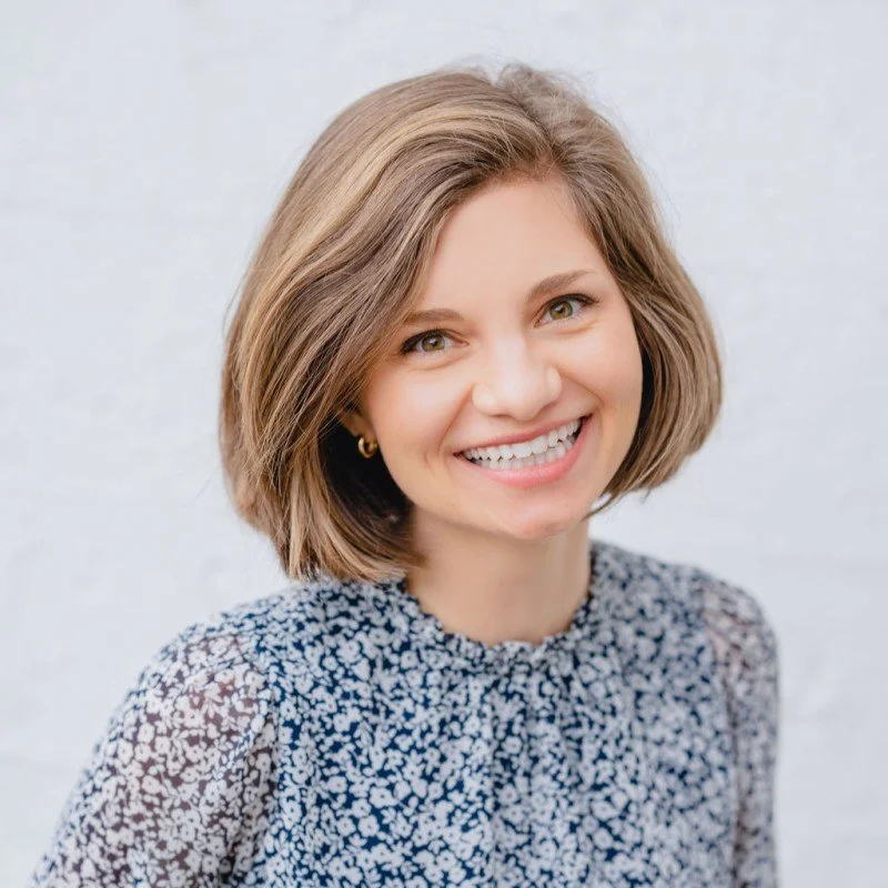 Smiling woman with short brown hair wearing a floral patterned blouse in front of a light background.