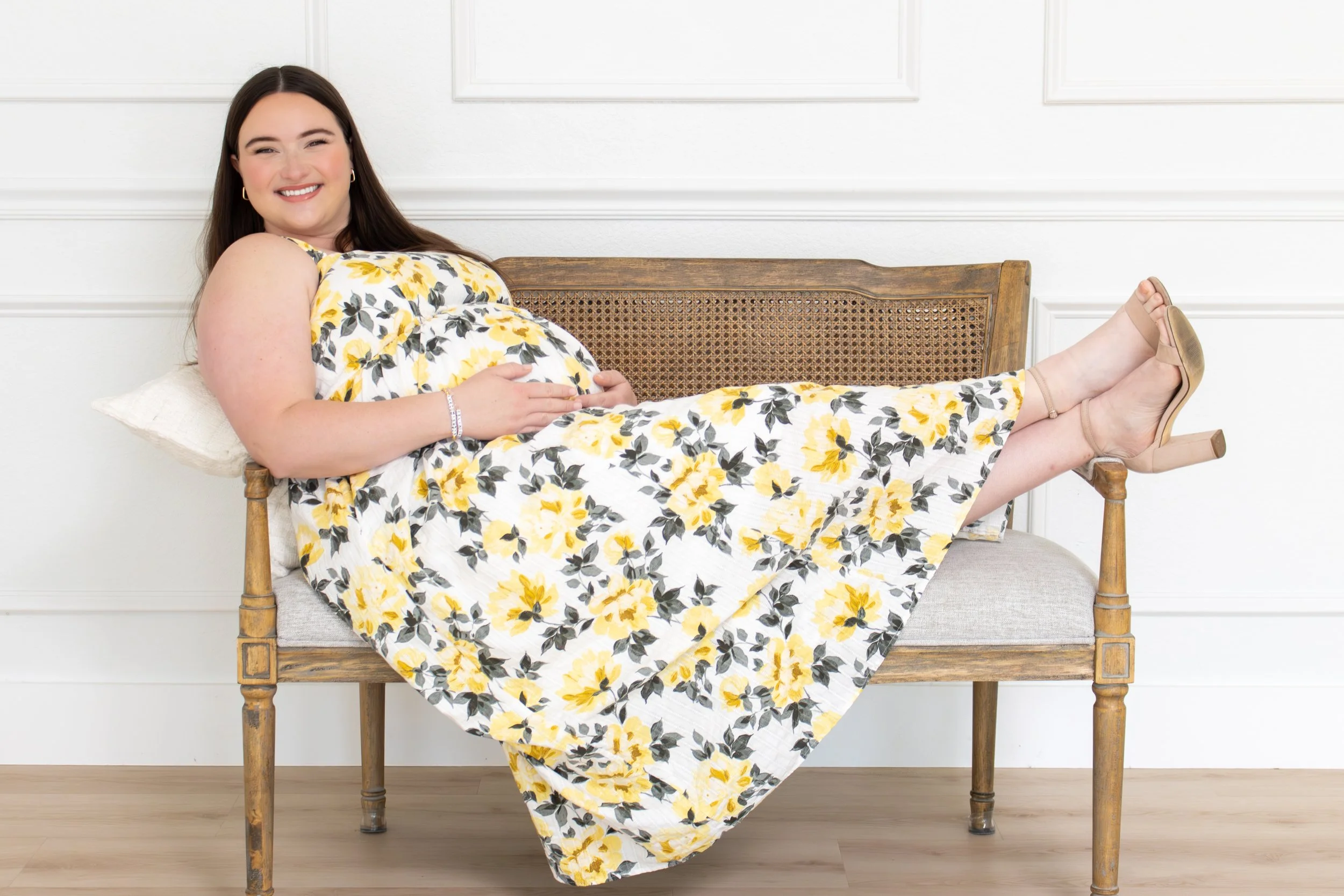 Pregnant woman lying on her back on a bed, smiling and resting her hand on her belly in a bright, airy room.