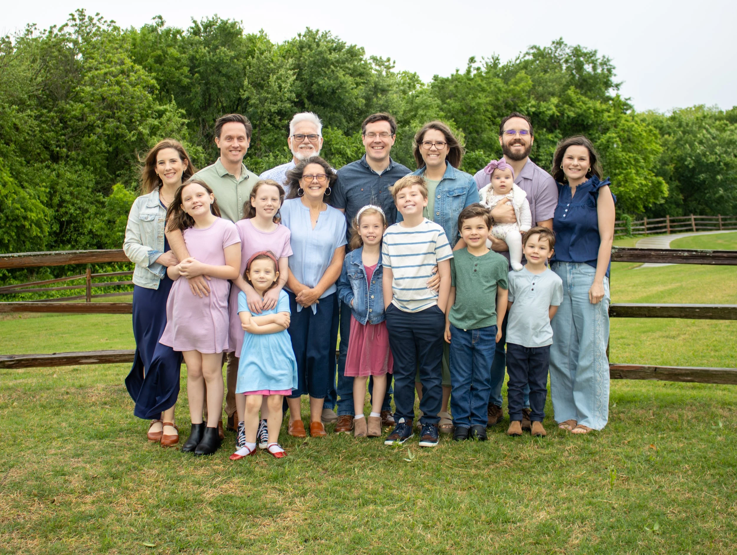 A smiling family of three sitting on a park bench outdoors with green trees in the background. The woman has curly hair and wears a white dress, the young boy wears a white shirt, and the man has a beard, dark hair, and wears a brown sweater.