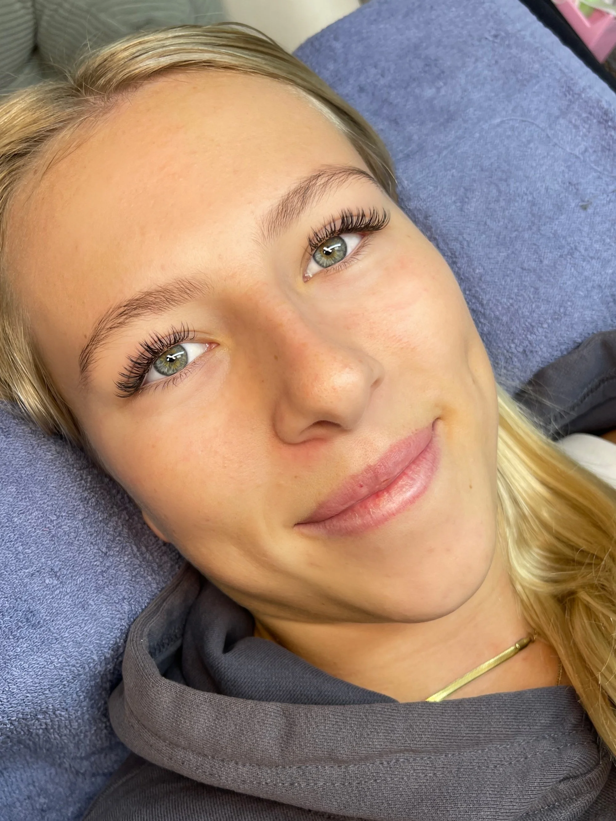Close-up of a young woman's face with light skin, green eyes, long eyelashes, and blonde hair. She is lying on a blue towel or blanket and smiling softly.
