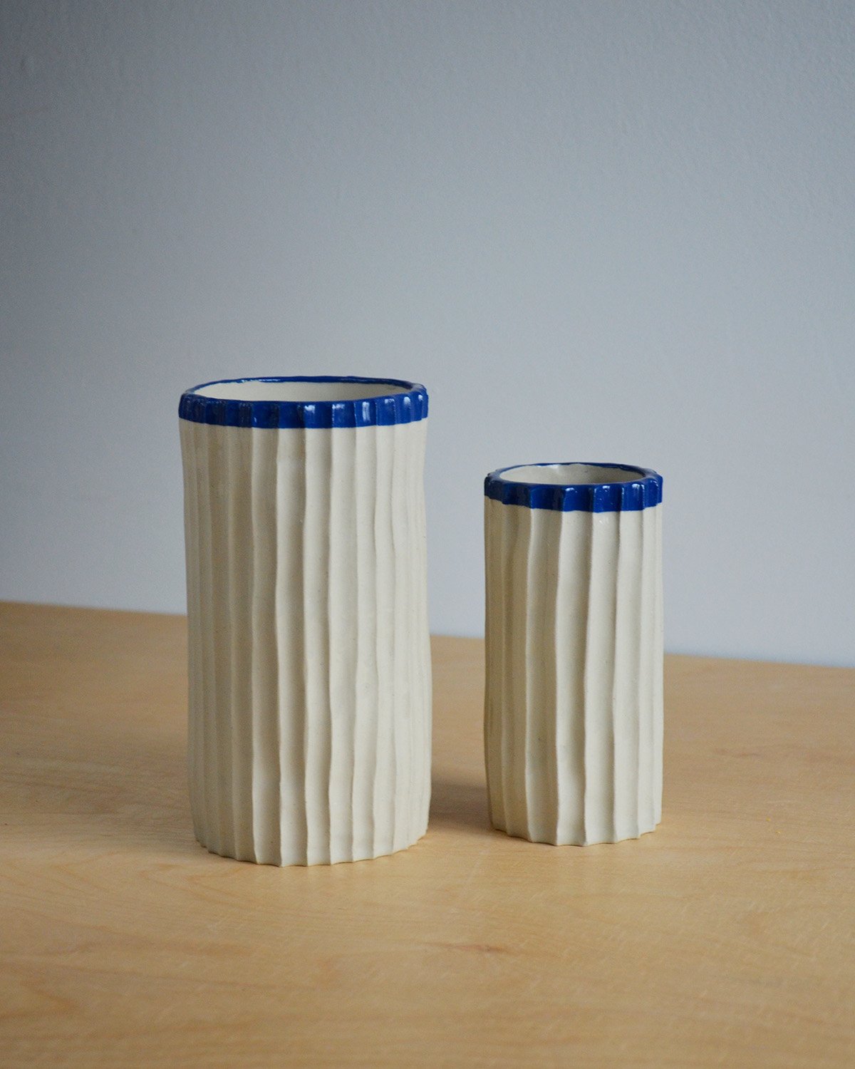 Set of two cream-white ceramic vases with a simple cylindrical design, grooved texture, and a glossy transparent glaze, accented by a bold blue rim—perfect for daily use or as a thoughtful gift.