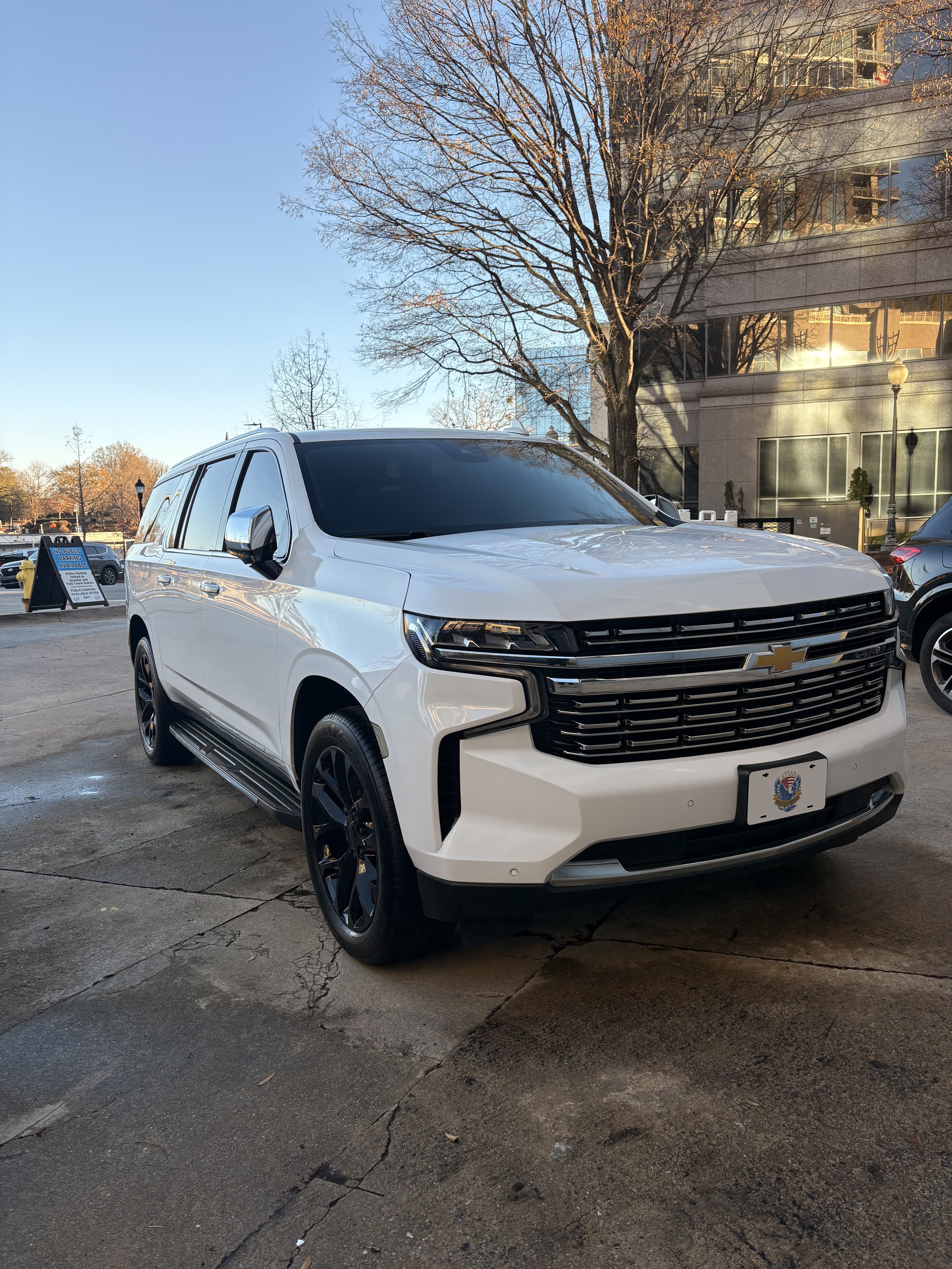 White Chevrolet SUV parked on a sidewalk in front of an office building with reflective windows, leafless trees, and a clear blue sky.