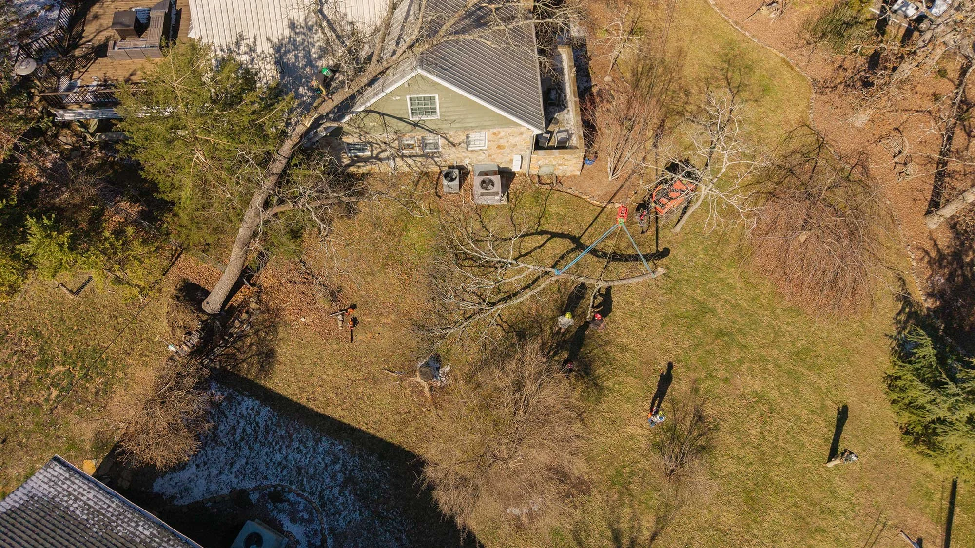 An aerial drone view showing a professional tree removal operation in a residential Chattanooga yard, featuring a large crane safely lowering a massive tree section near a house while ground crews manage the debris.