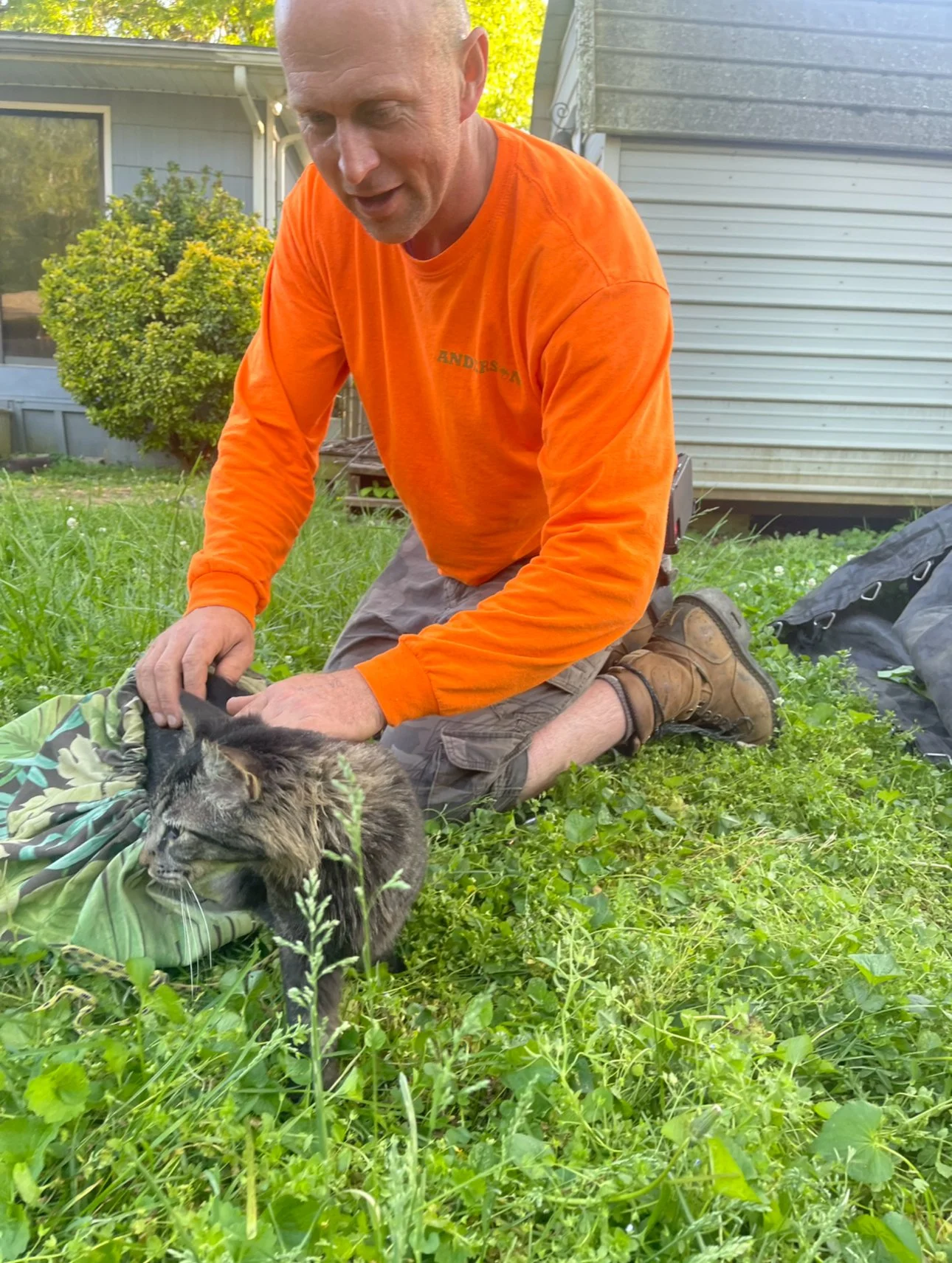 Jon Cable from Anderson Gutter & Tree kneeling on grass, wearing an orange long-sleeve shirt and brown shorts, petting a gray and black cat.