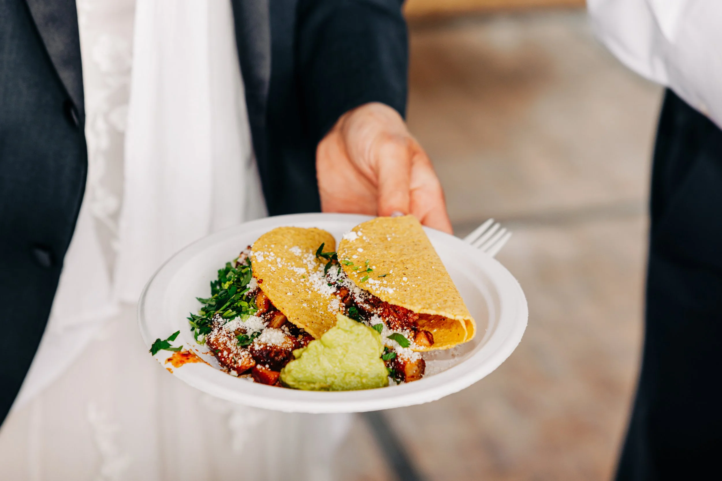 Hard shell tacos with guacamole and cilantro on a plate held by a wedding guest.