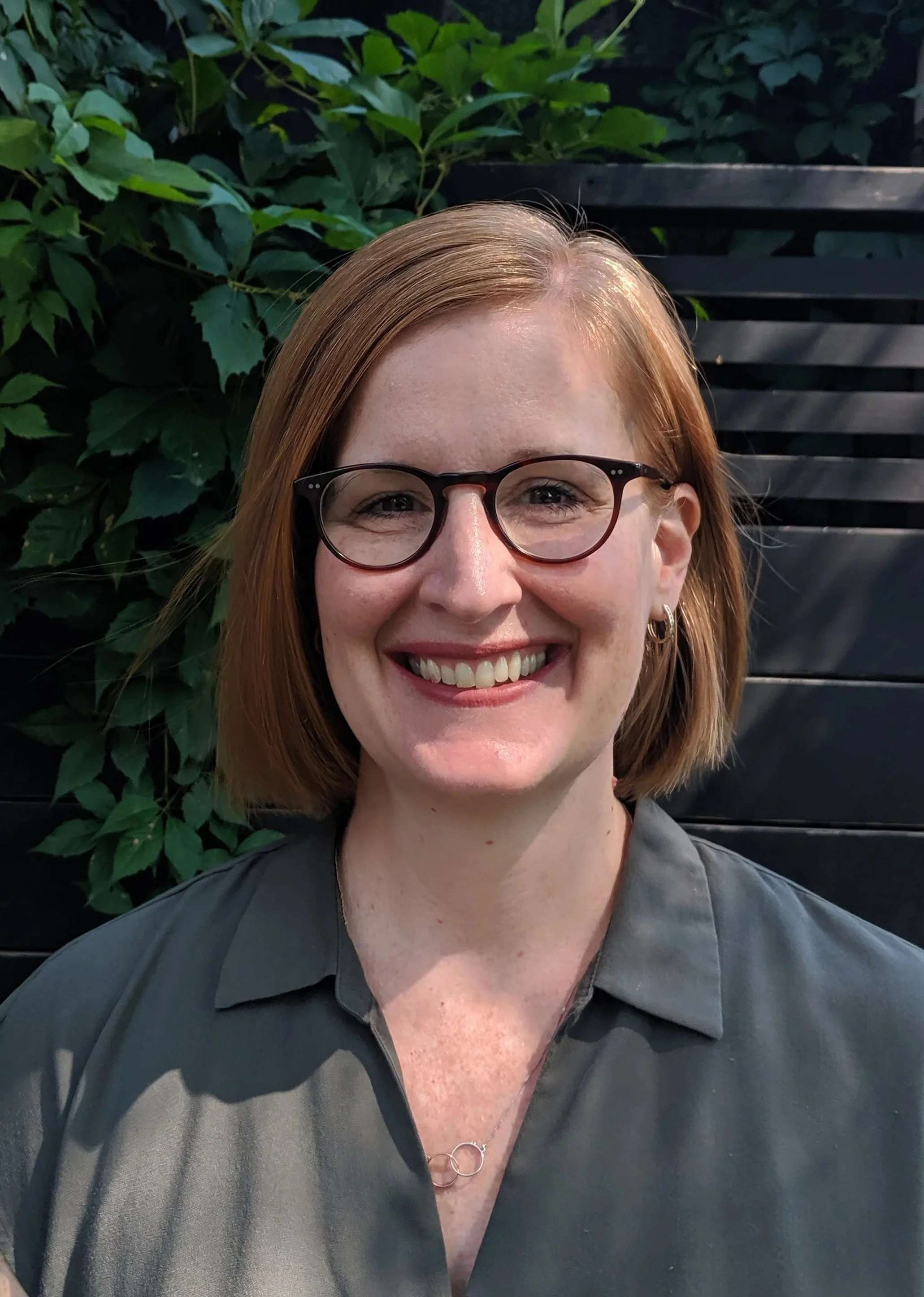A woman with shoulder-length red hair, wearing glasses and a dark gray shirt, is smiling outdoors in front of a background of green leaves and a black wooden fence.