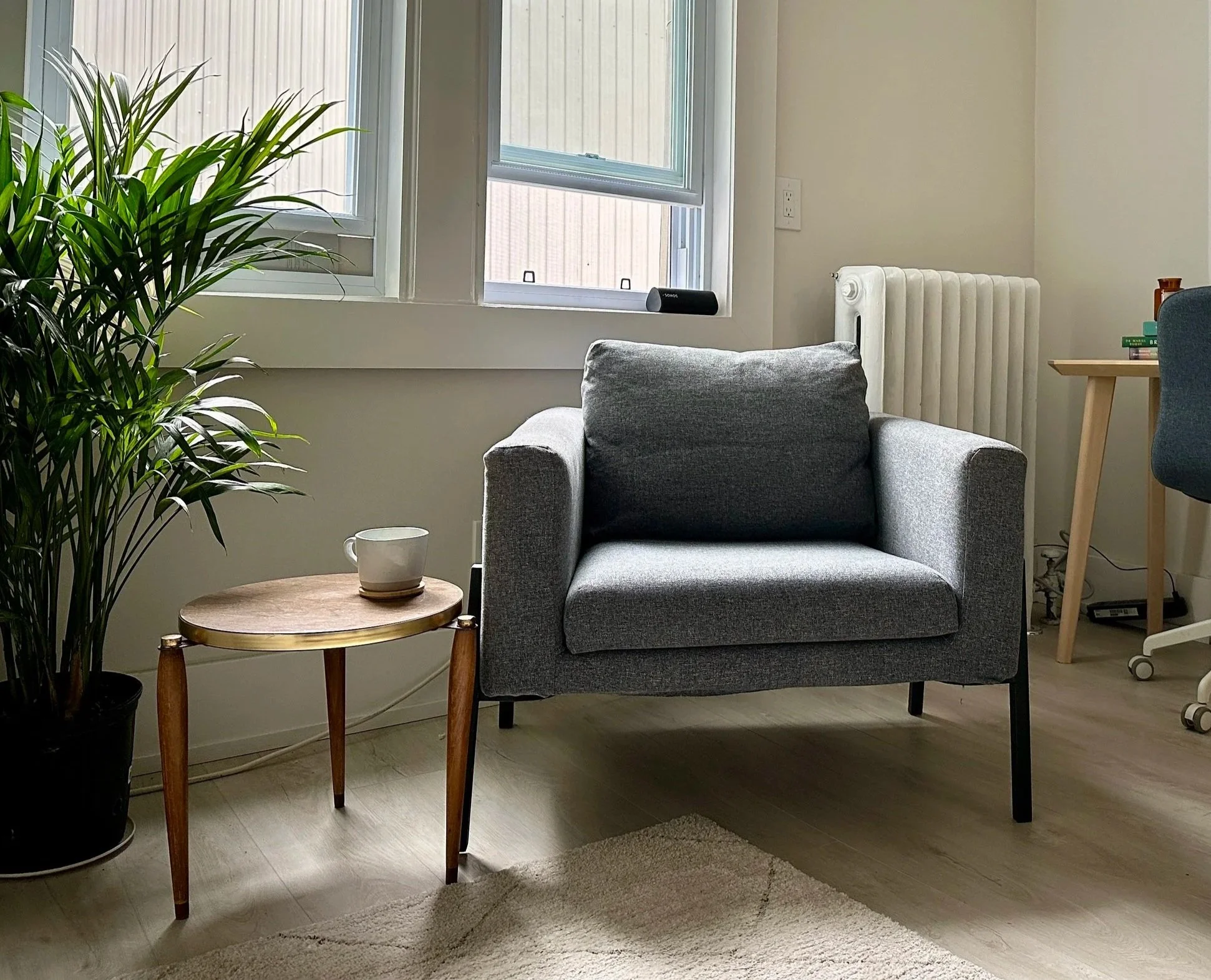 A cozy living room corner with a gray armchair, a small wooden side table with a white mug, a potted plant, a window with closed blinds, a radiator, and a corner of a desk with a blue chair.