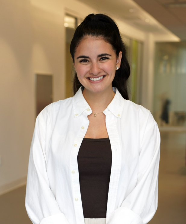 Smiling woman with dark hair in a ponytail, wearing a white button-up shirt over a black top, standing in an indoor office environment.