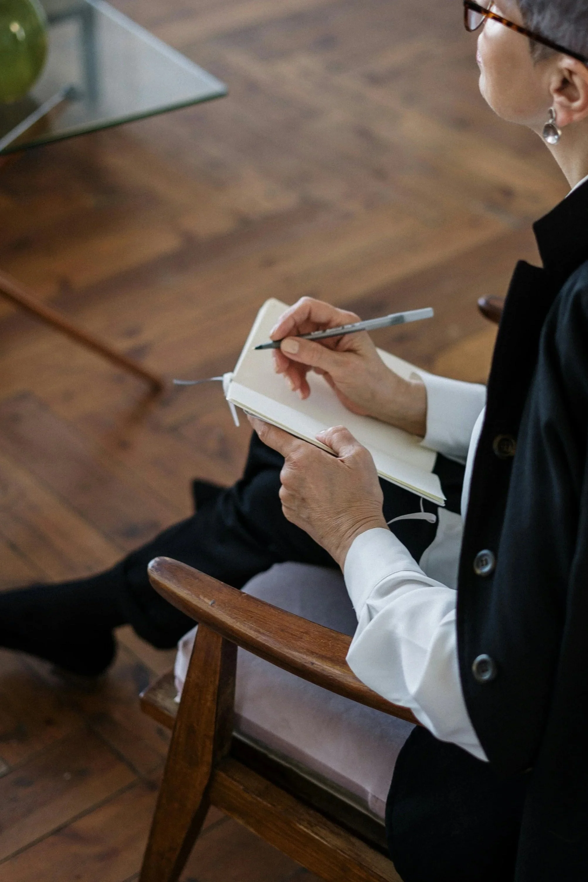 A woman is sitting on a wooden chair, writing in a notebook with a pen, in a room with wooden flooring.