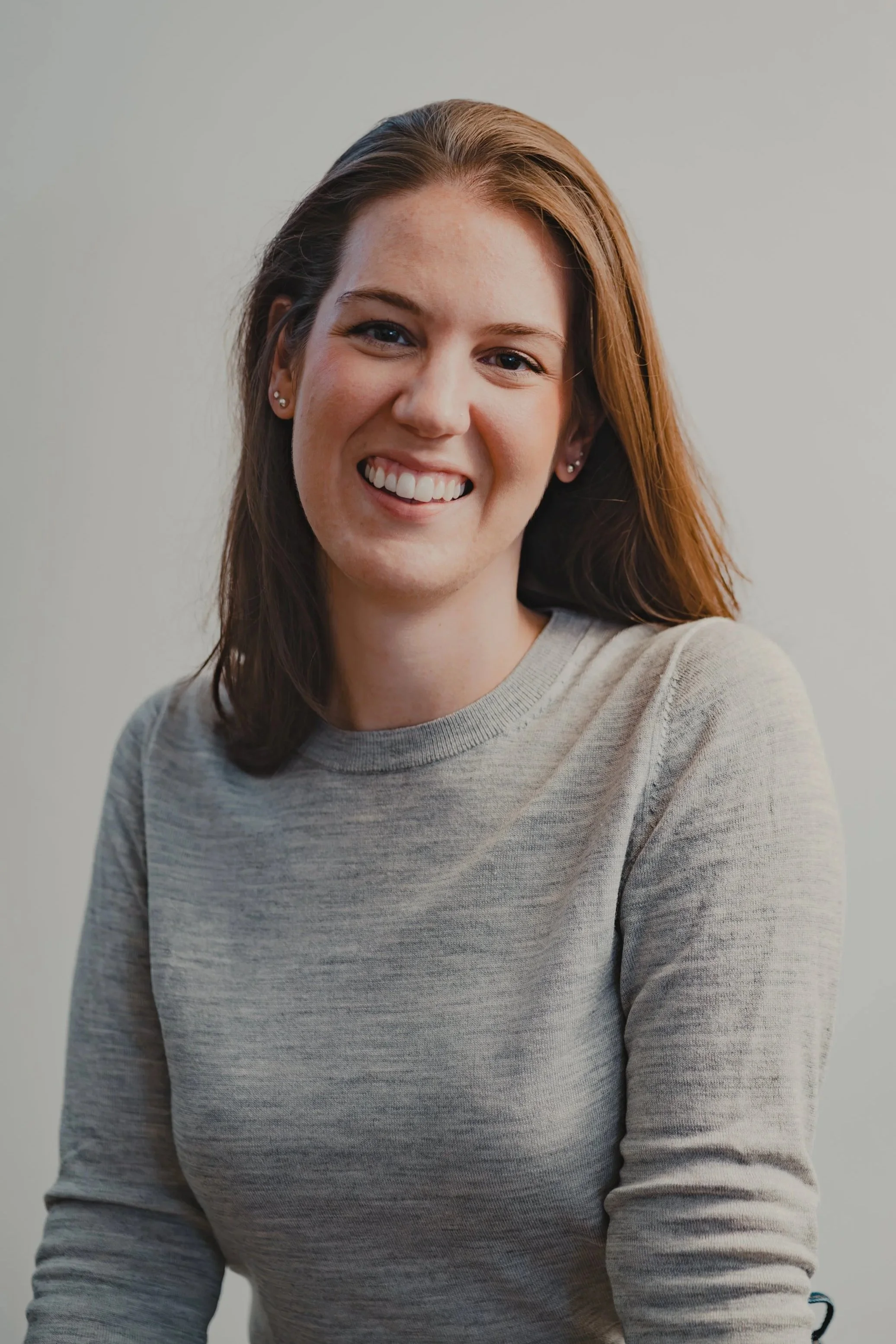 A young woman with red hair and earrings smiling in front of a plain light-colored background.